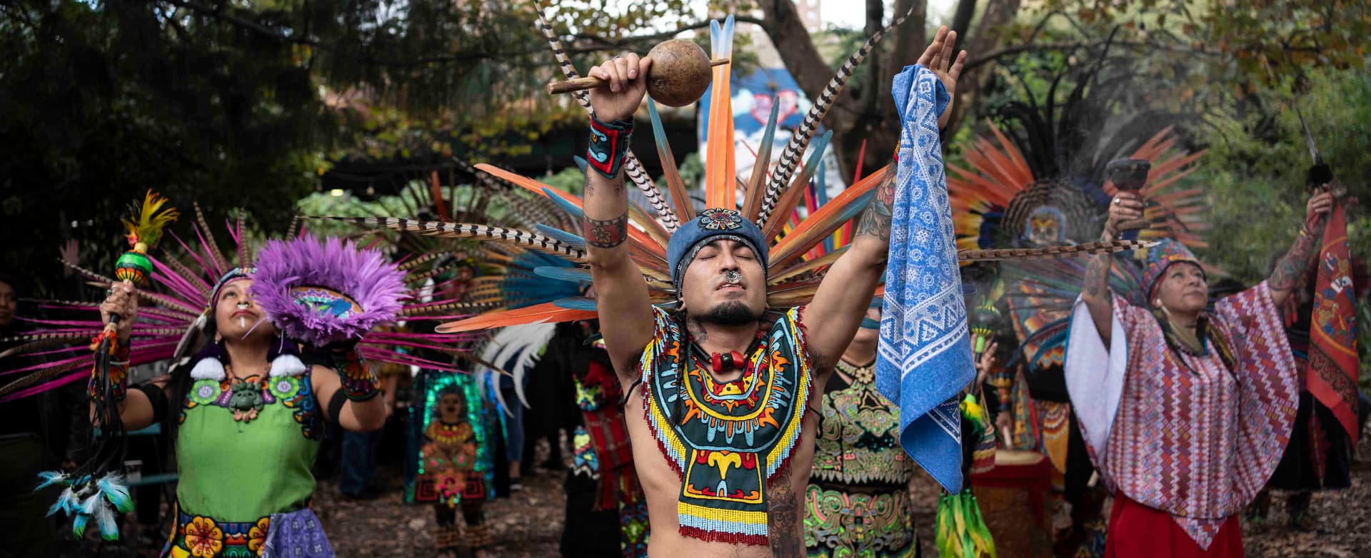 Group performs ceremonial dance in colorful feathered regalia, arms raised outdoors.