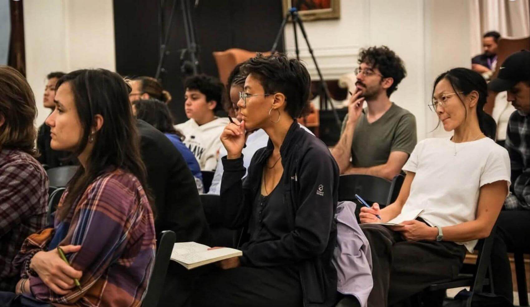Two women sit in the audience, listening and taking notes, with several people behind them.
