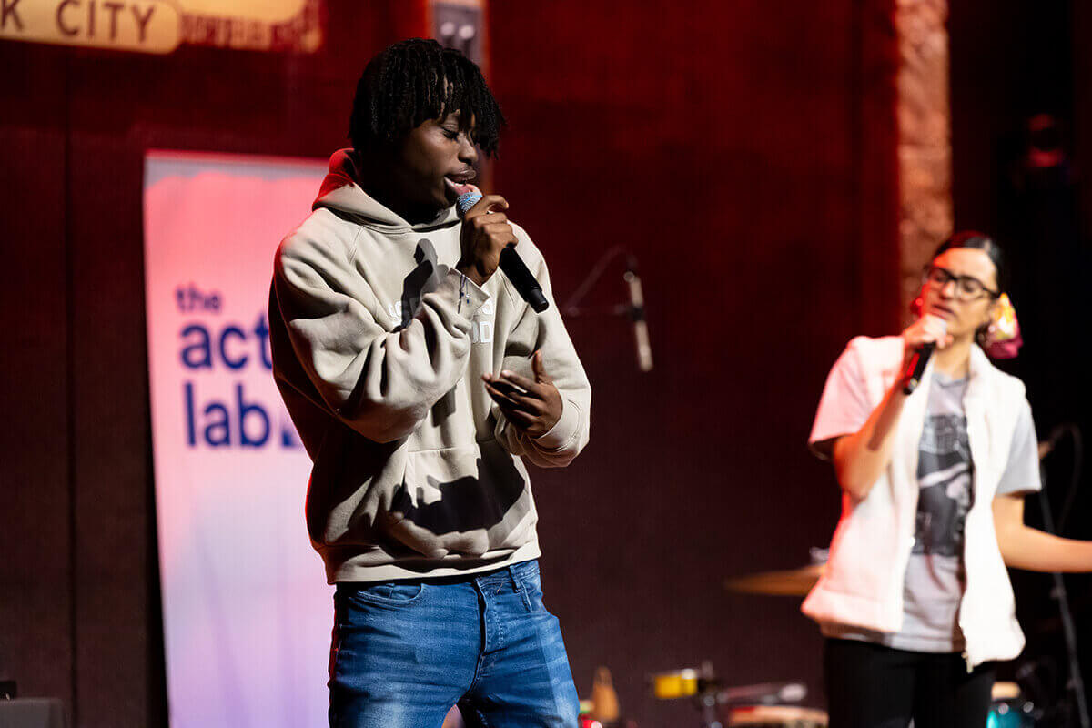 Group of people on stage at City Winery New York, one person speaking into a microphone and raising hand, others clapping or holding microphones.