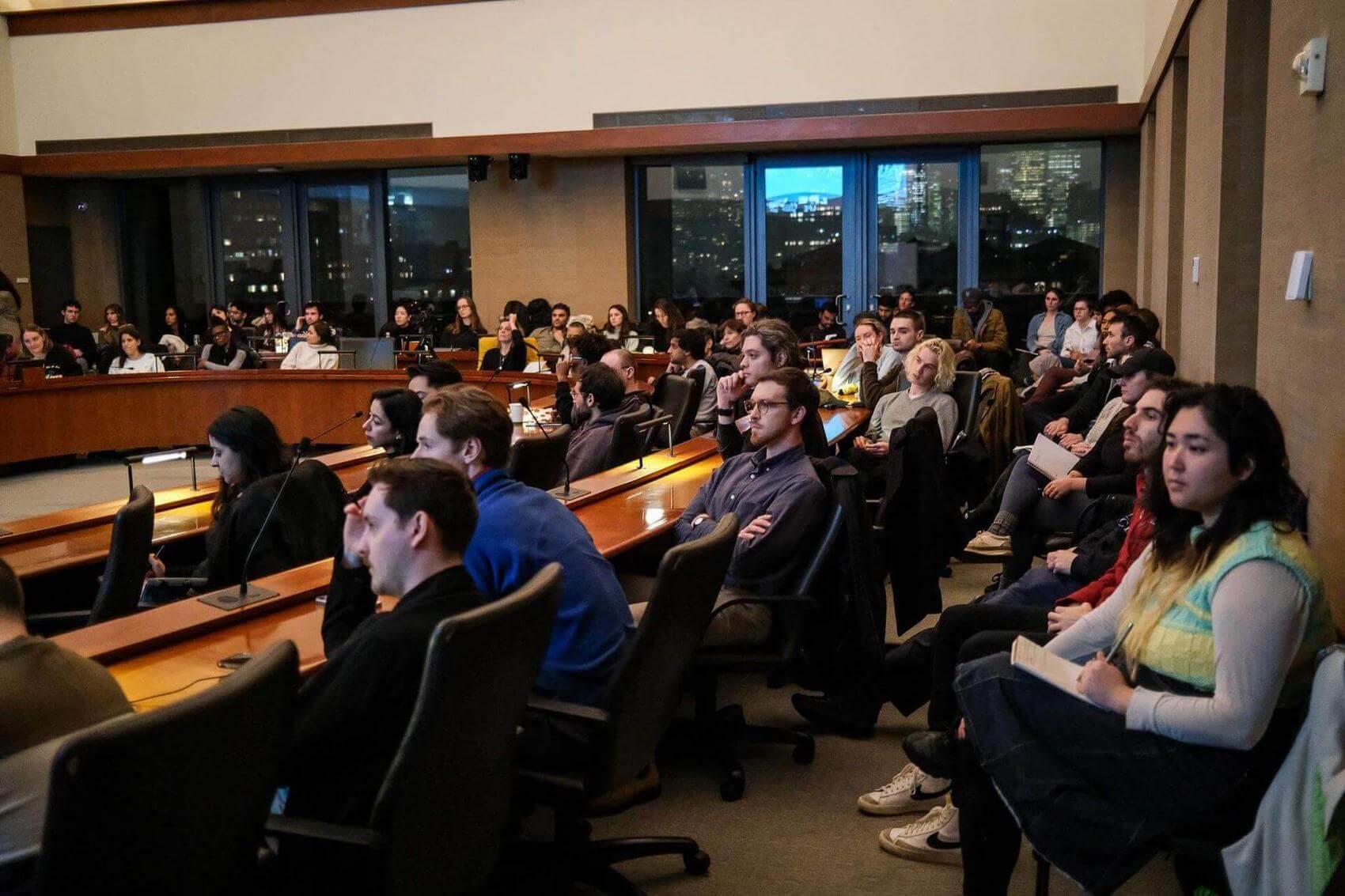 Two people seated at a boardroom table; one gestures while speaking, the other listens.