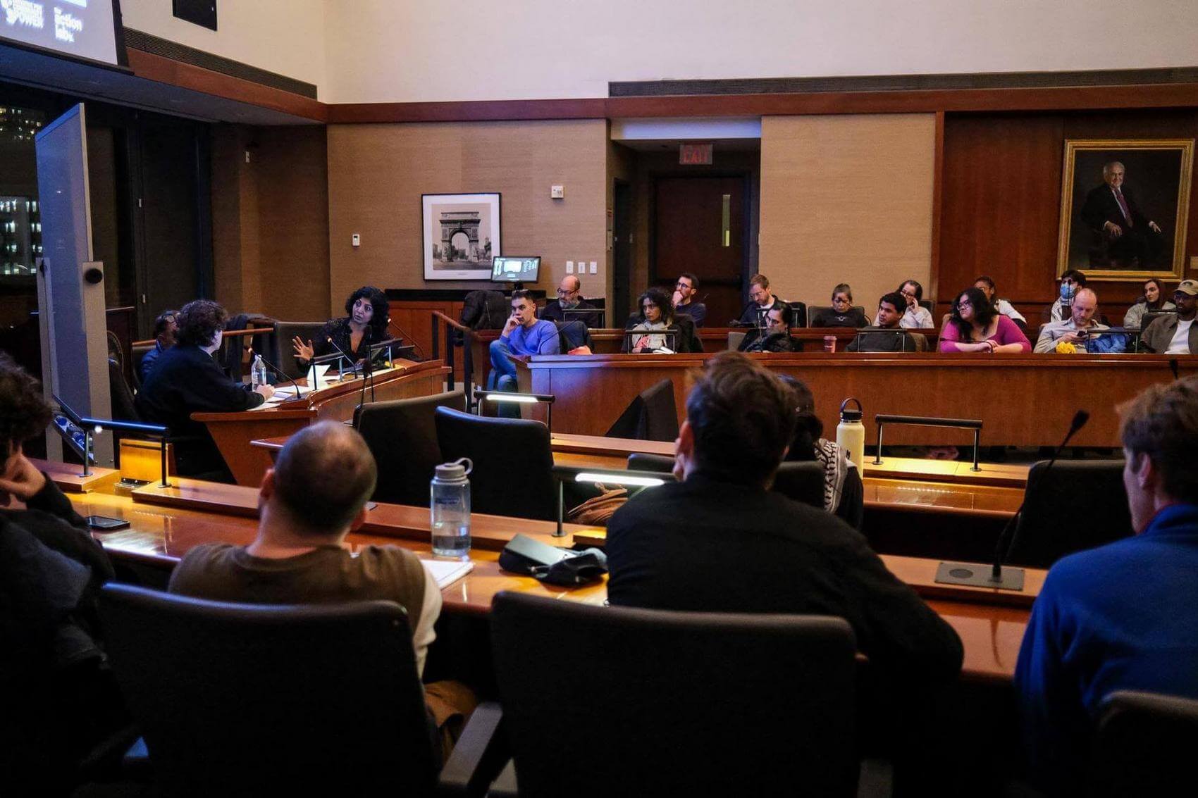 A group of people sitting in a conference room, some listening attentively while one writes notes.