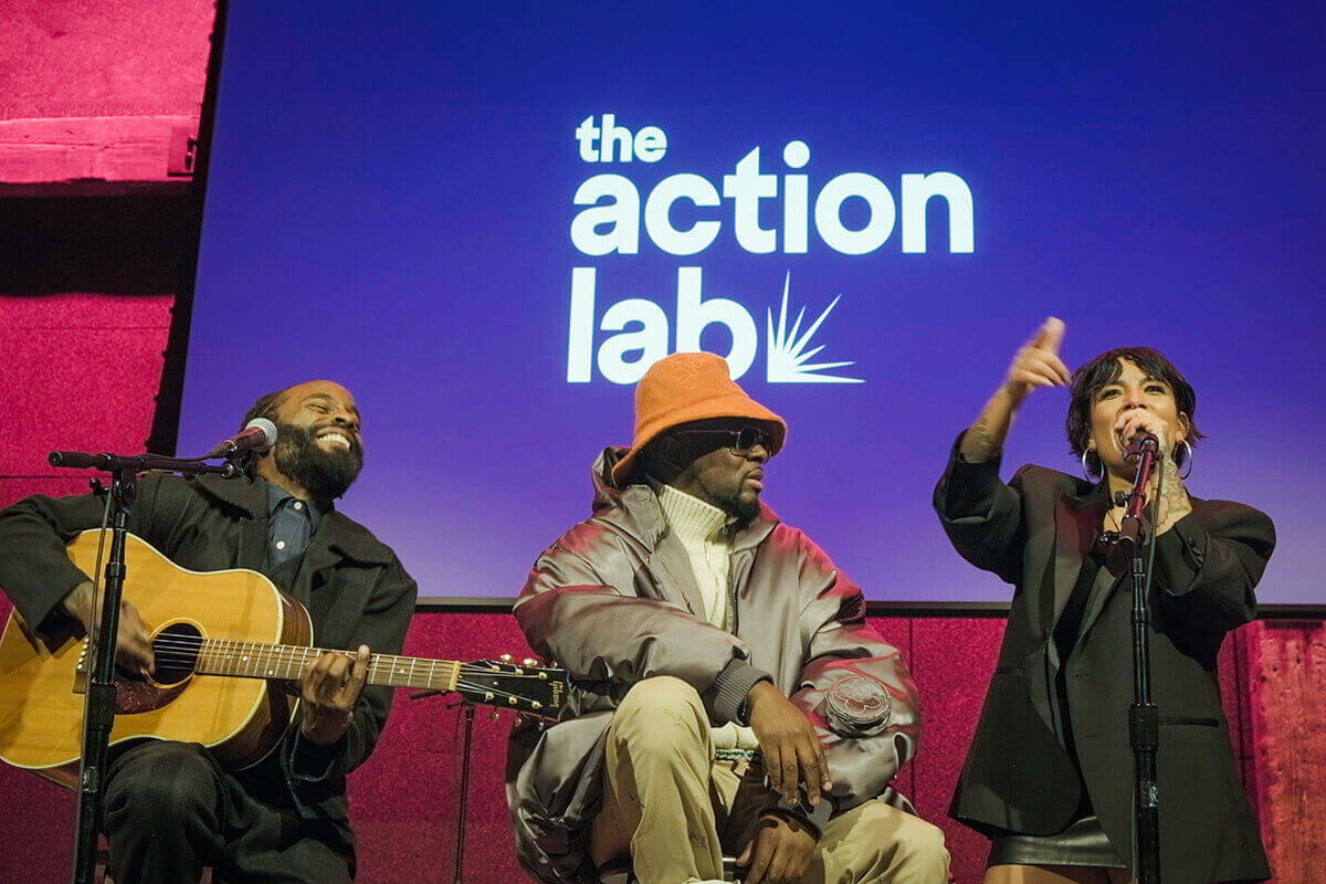 Three people performing on stage in front of a screen with "the action lab" text, one playing guitar, one sitting, one speaking into a microphone.