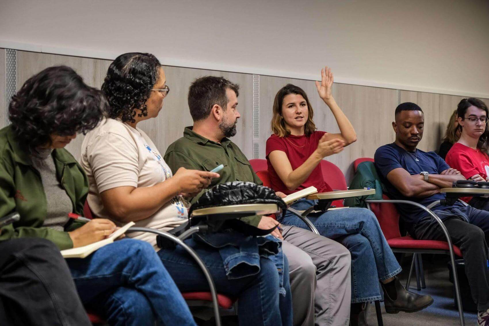 A group of six people seated in a classroom; one person is speaking with raised hand.