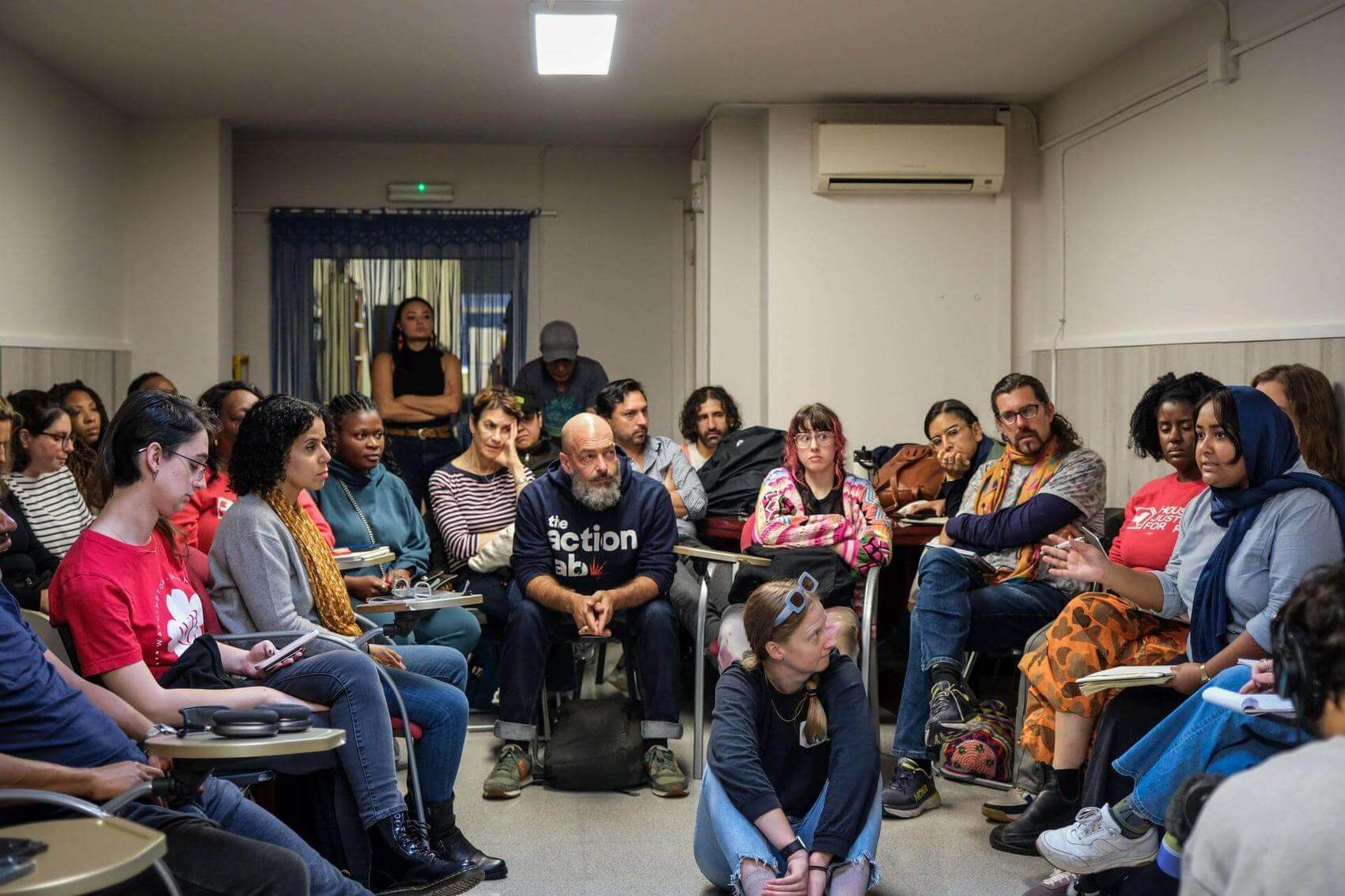 A diverse group of people seated in a room discussing, some taking notes.