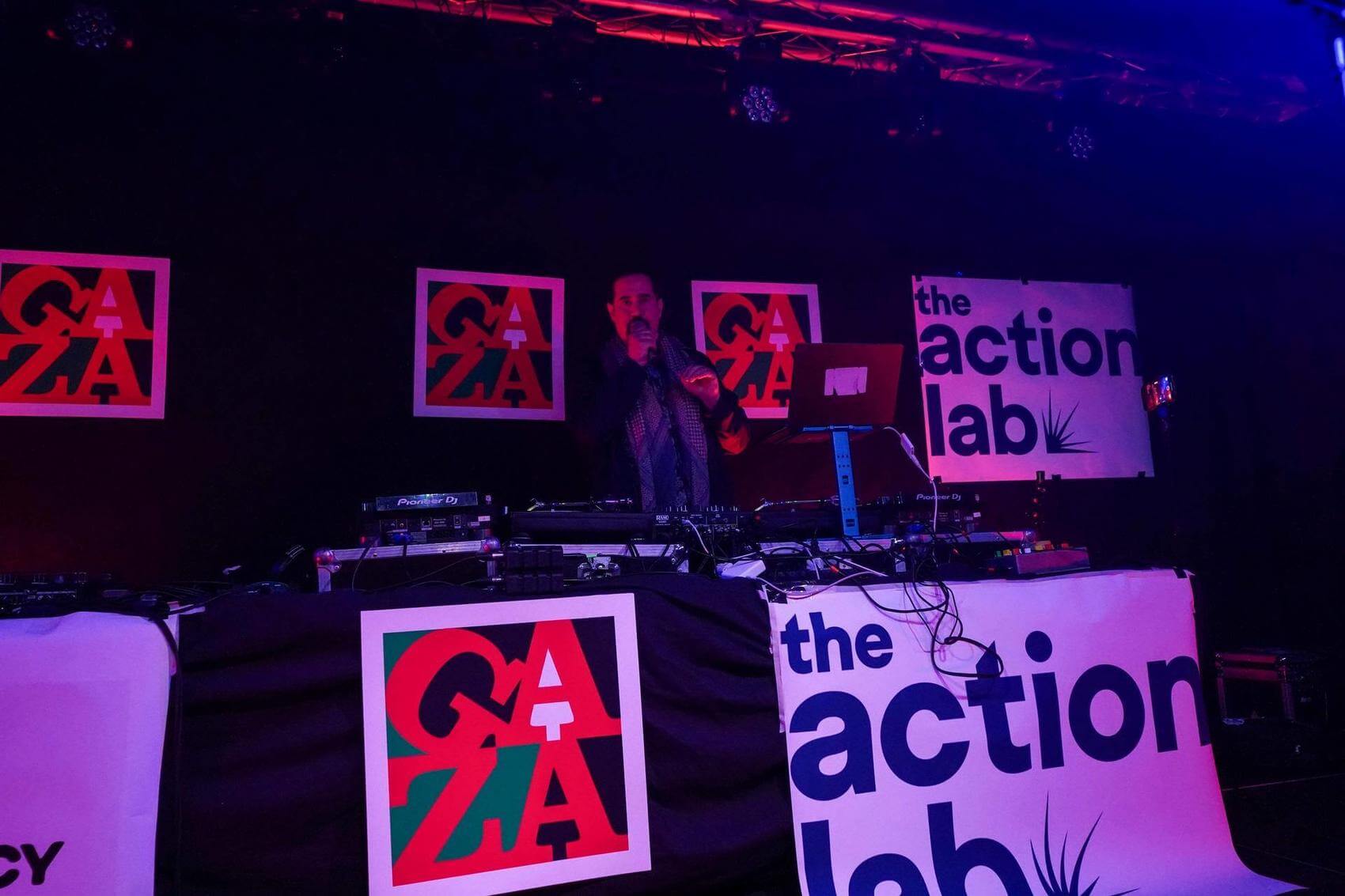 Two performers rapping into microphones on stage with DJ equipment in the background.