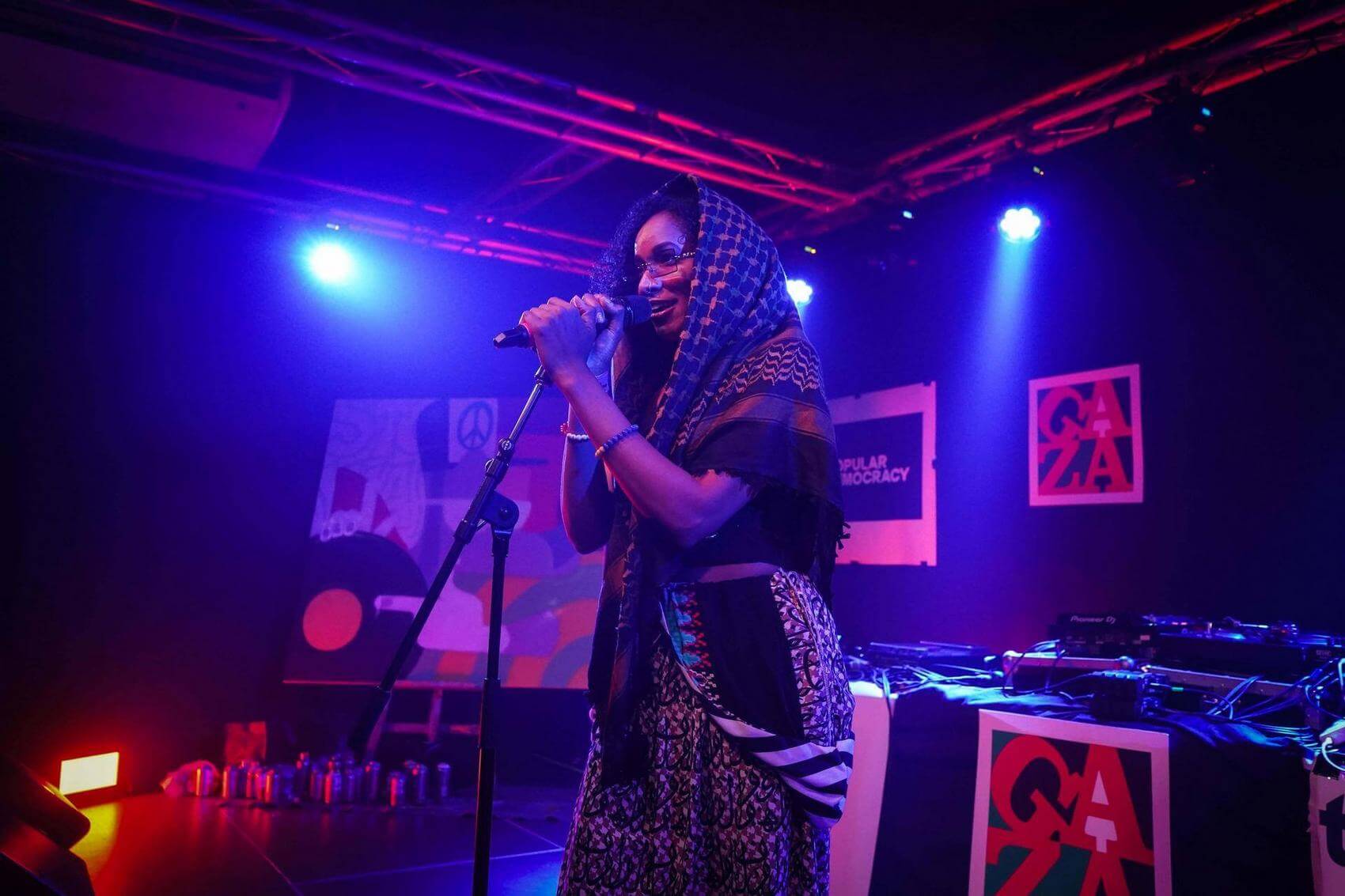 Two people sit at a table with electronic music equipment, one using a smartphone. Bags and water bottles are visible in the background.