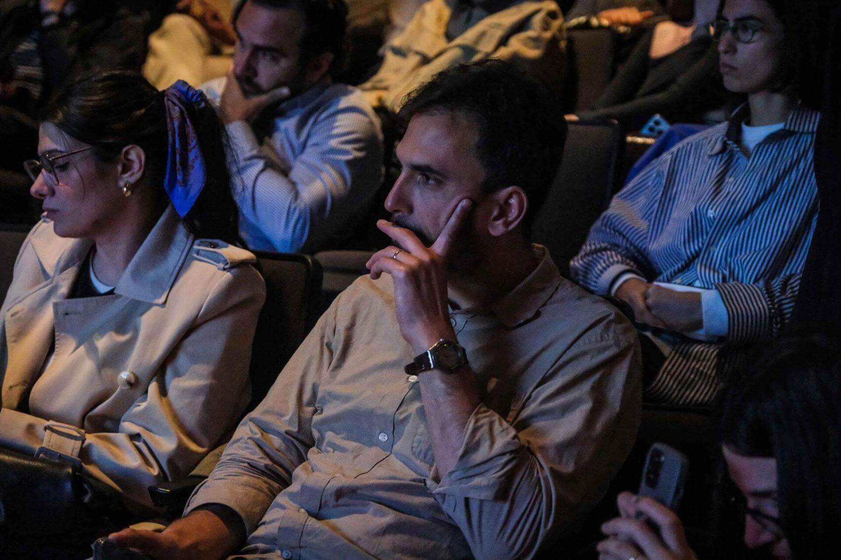 Audience members watching a presentation, with focused expressions seated in a dimly lit space.