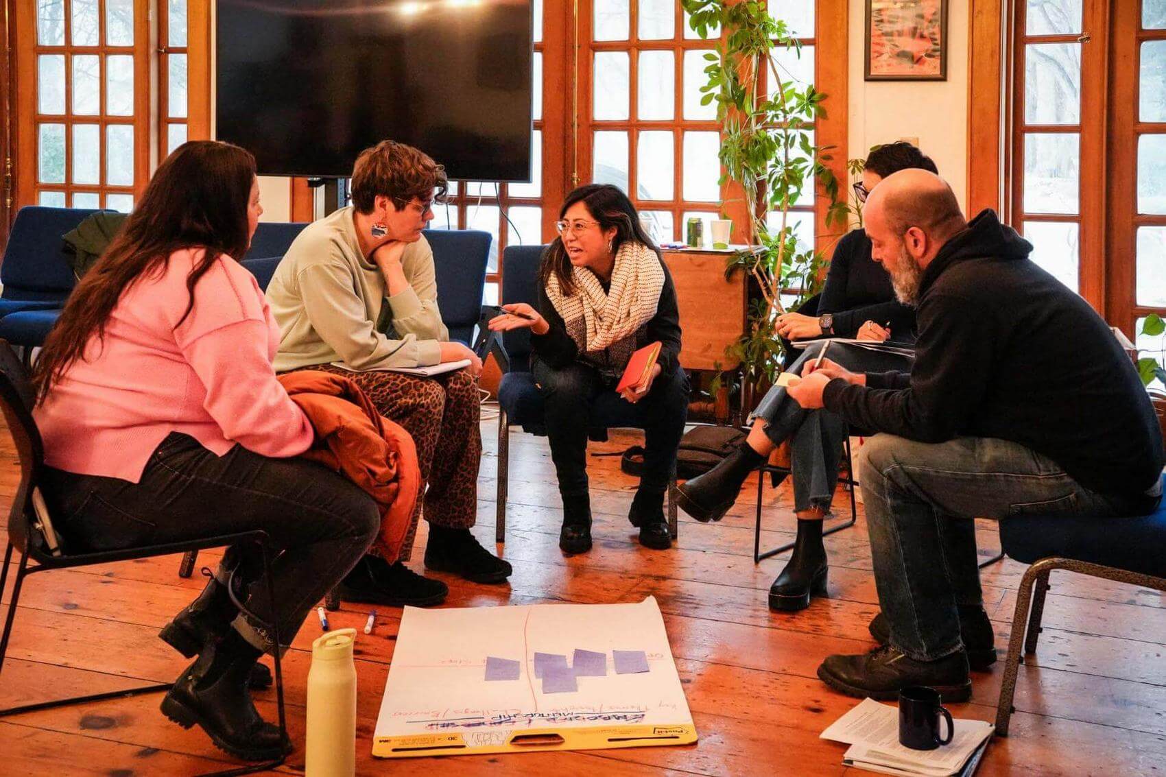 Five people seated in a circle having a discussion with notes and papers on the floor.