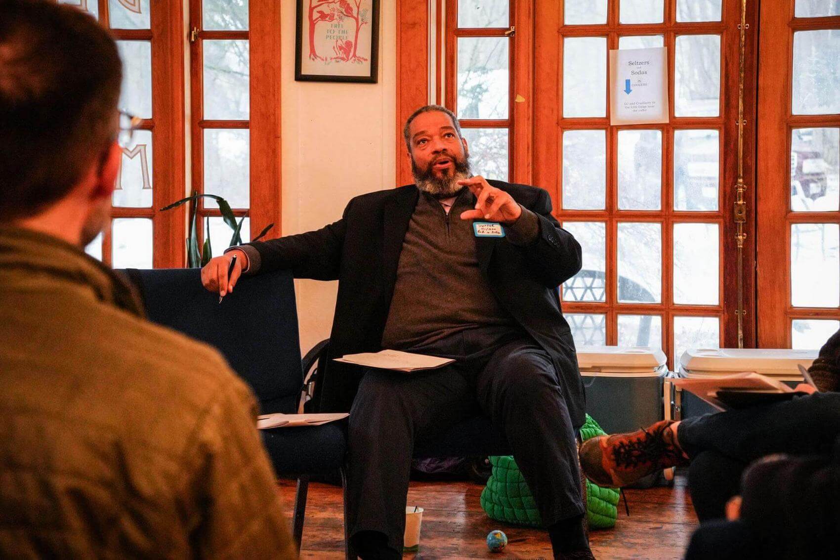 Man seated and speaking to a group, with a notebook and pen in hand, in a room with windows.