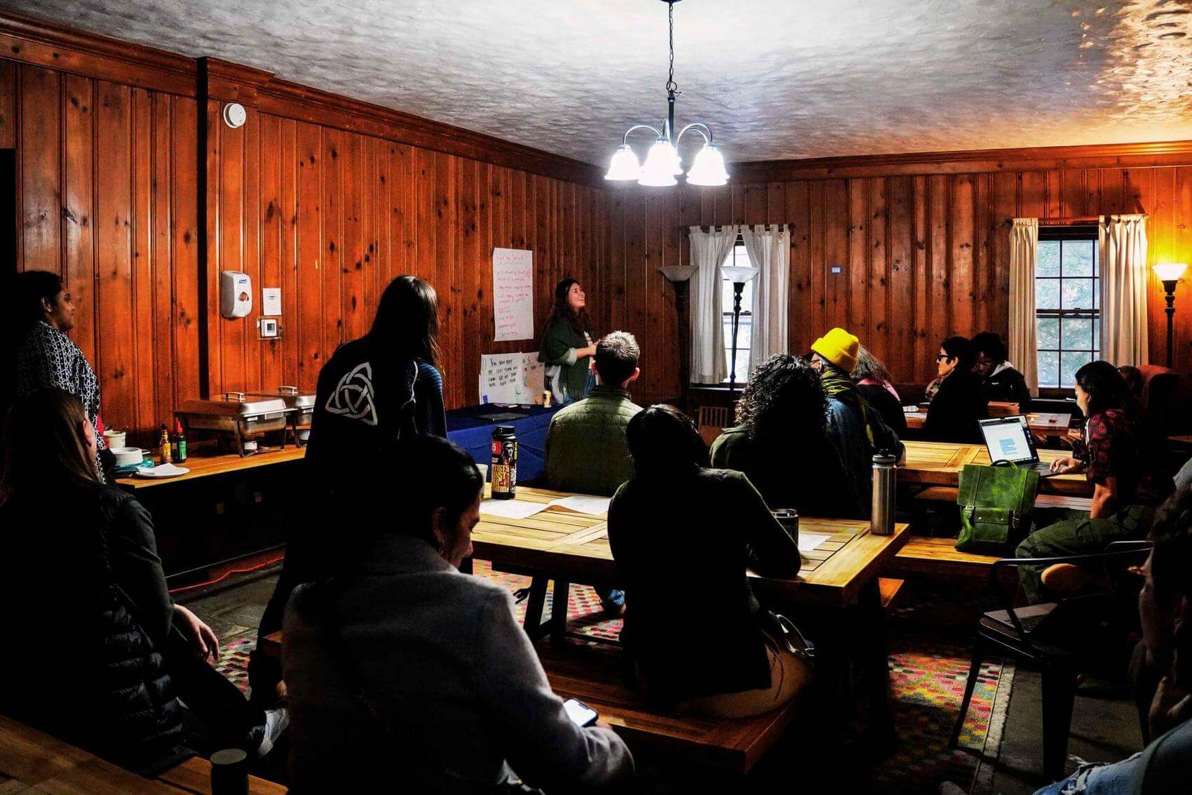 A group meeting in a room with large windows, a man speaking while others listen.