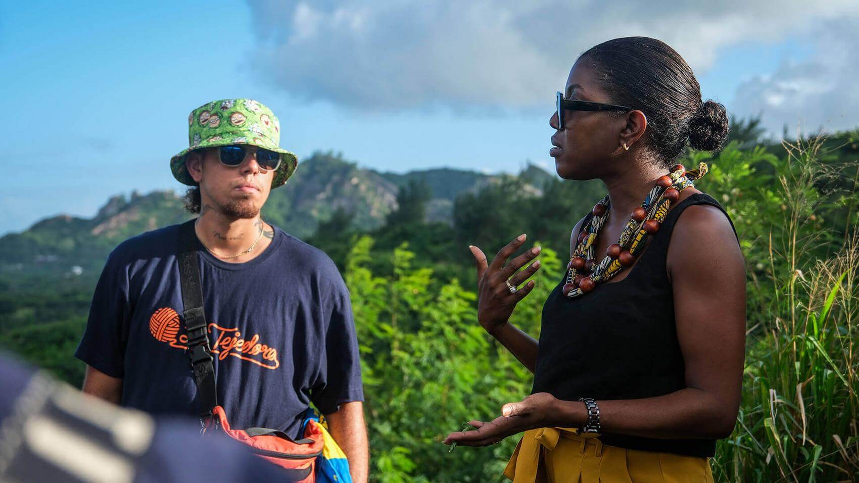 A woman gestures as she speaks to a man against a green mountainous backdrop.