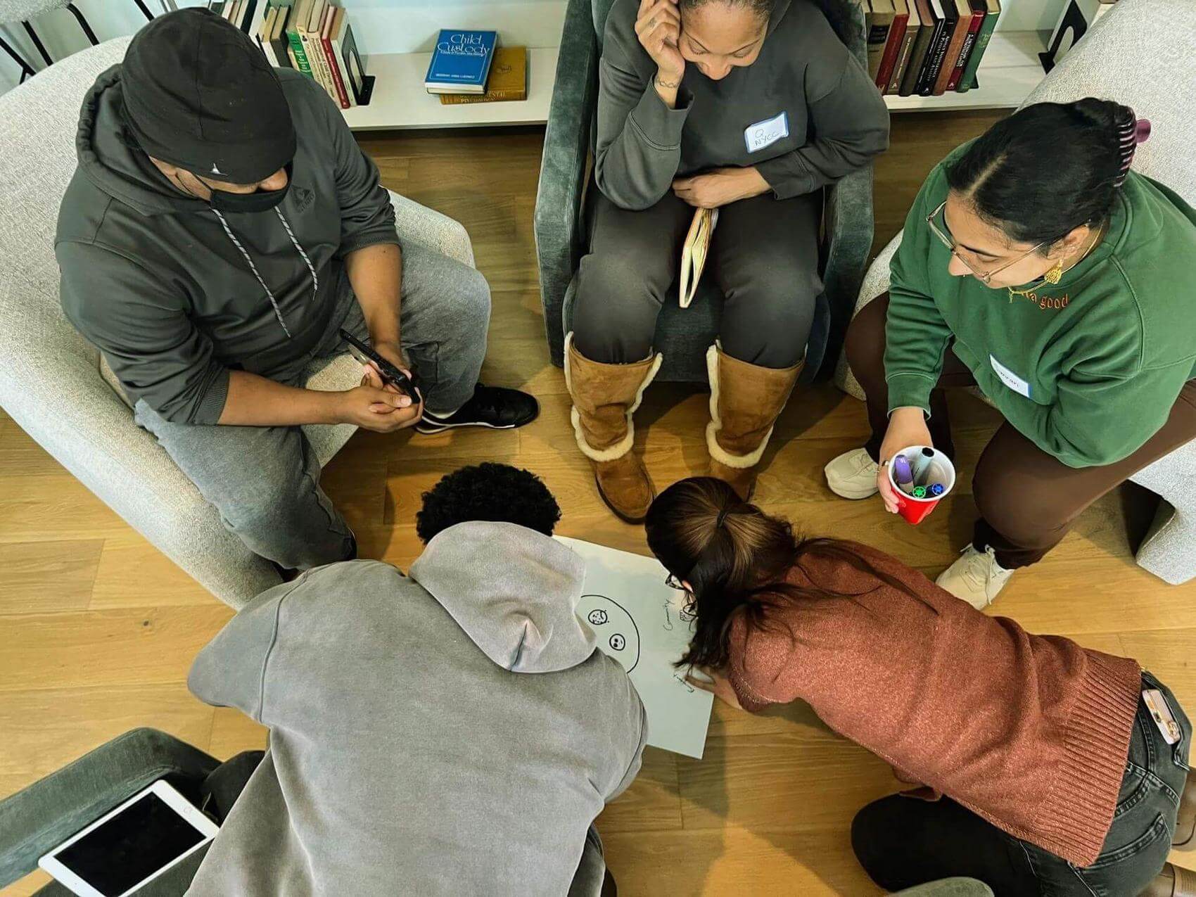 Six people gather around a large sheet of paper on the floor, engaging in a collaborative activity.