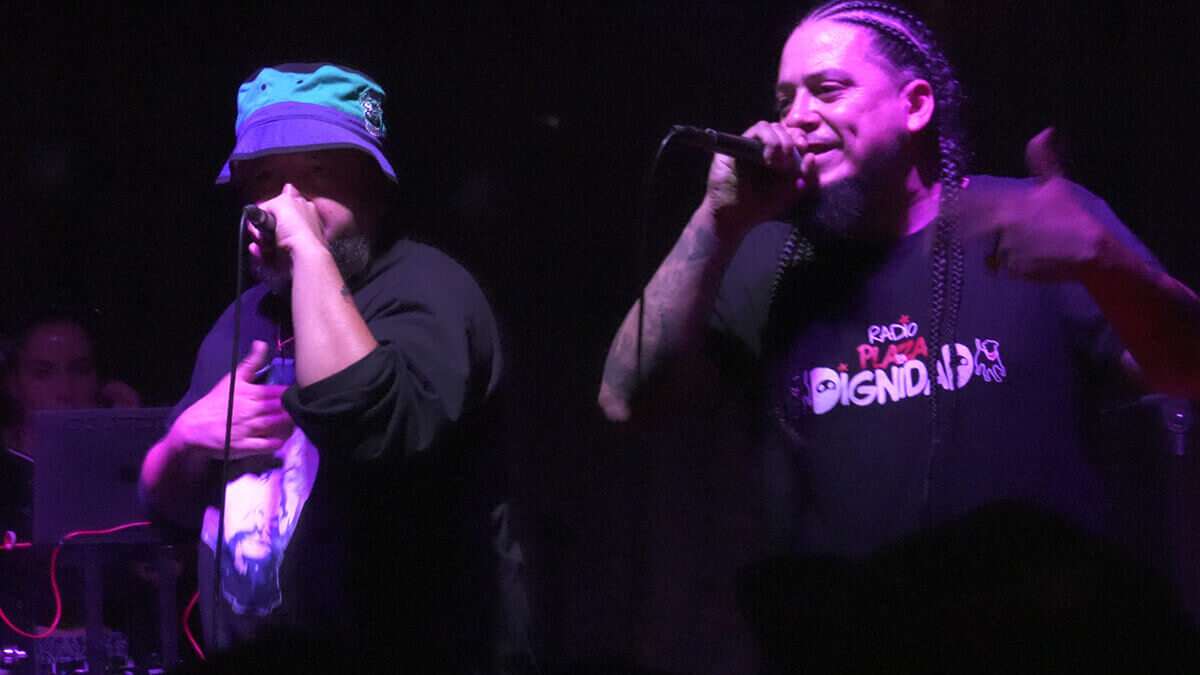 Person holding a microphone performing on stage with a DJ using a laptop behind them.