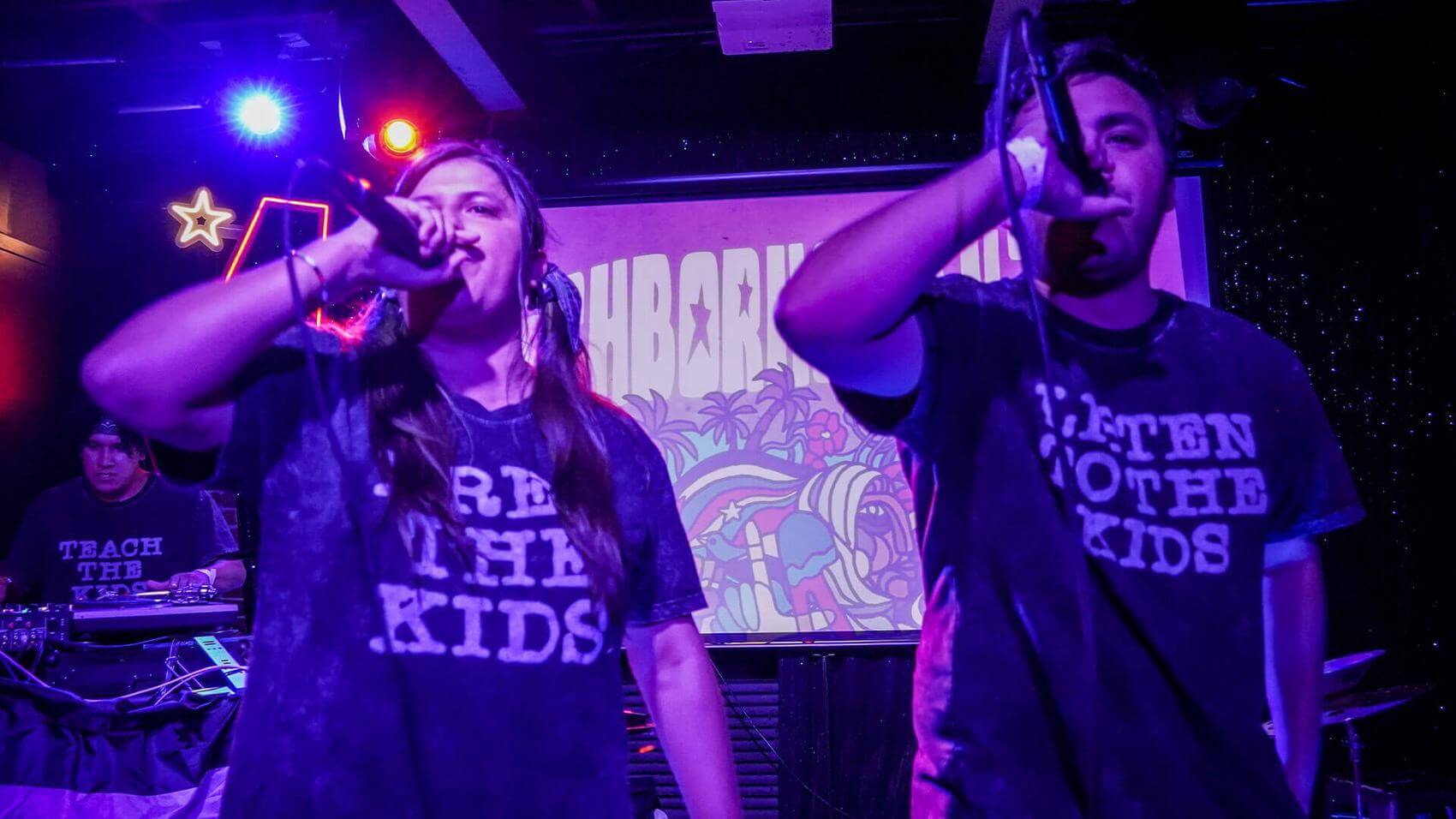 Woman singing into a microphone while holding a ukulele on a stage with purple lighting.
