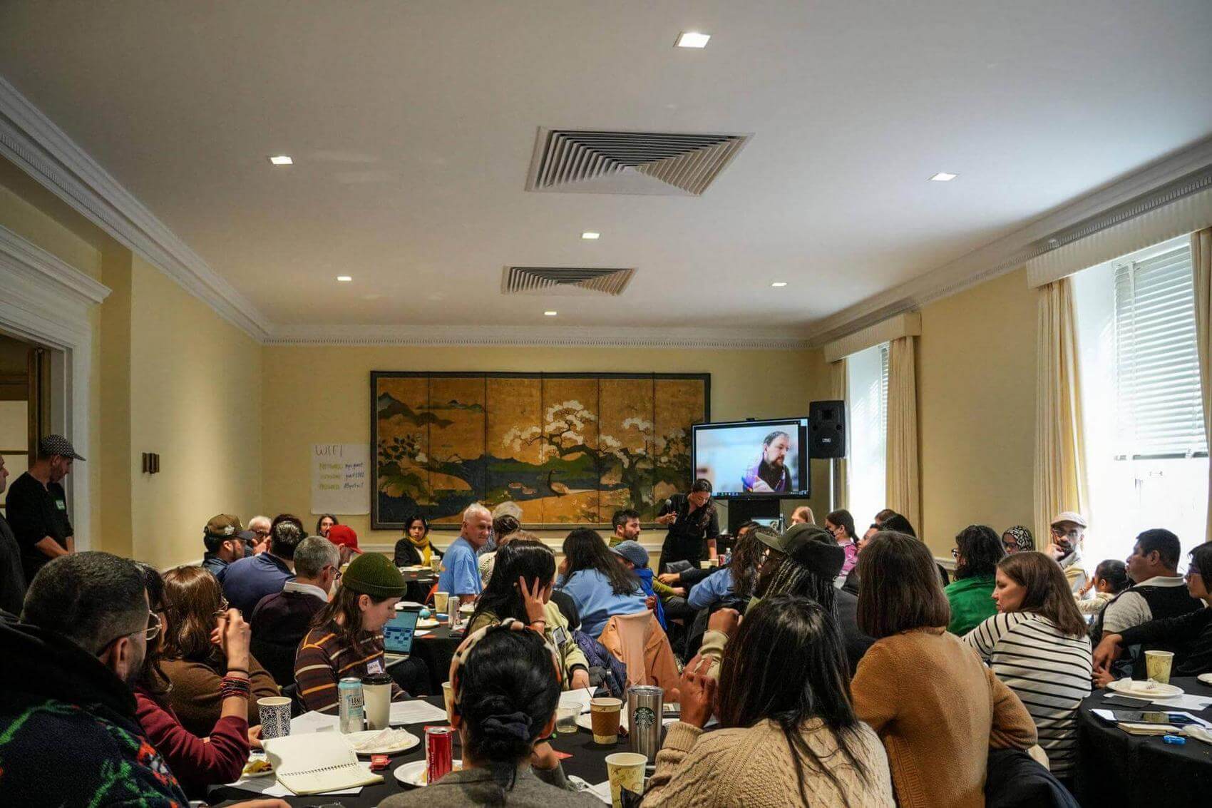 Group discussion in a large room with tables, a projector display, and a mural.
