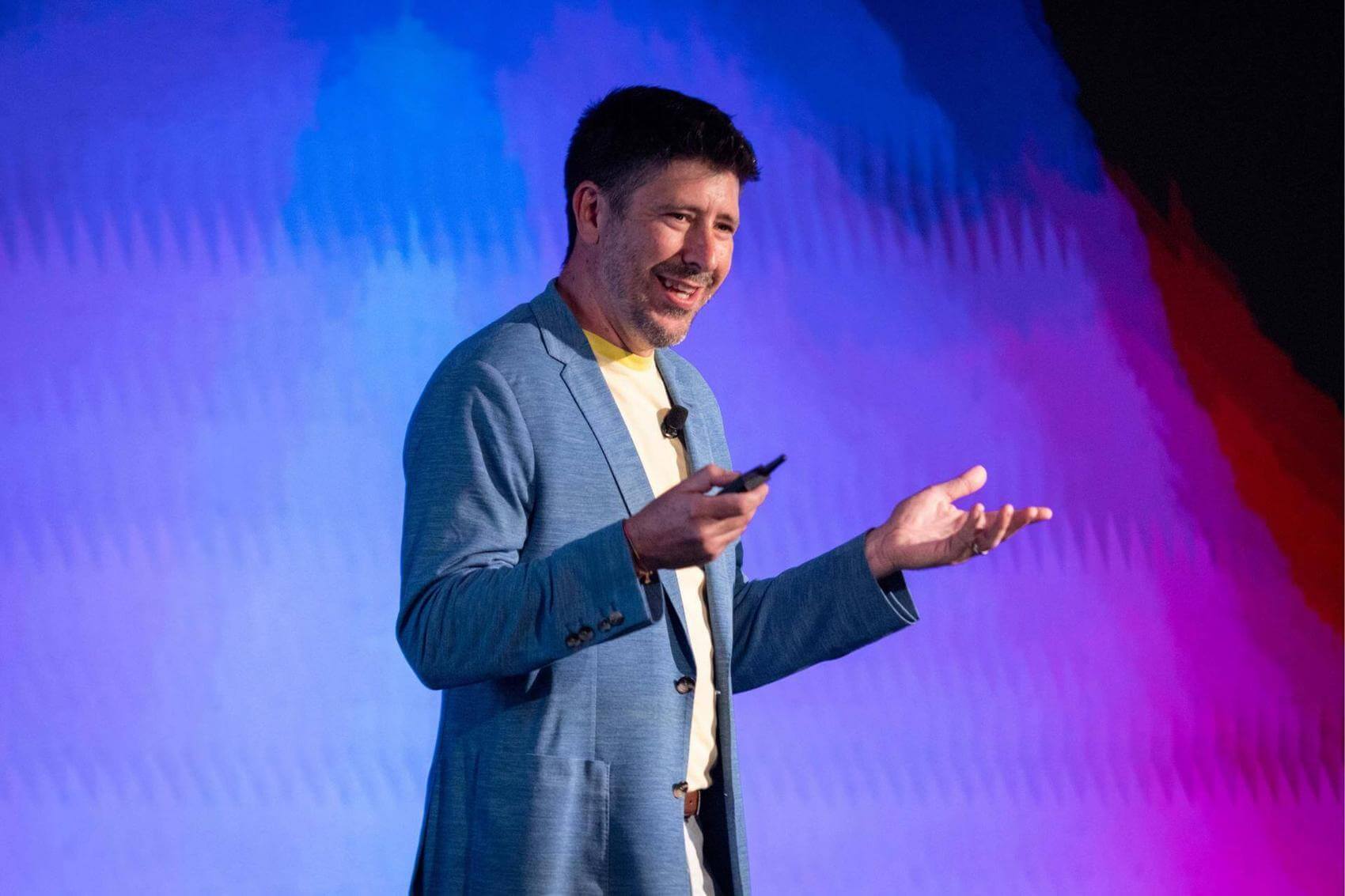 Woman speaking into a microphone in a gathering, with people in the background.