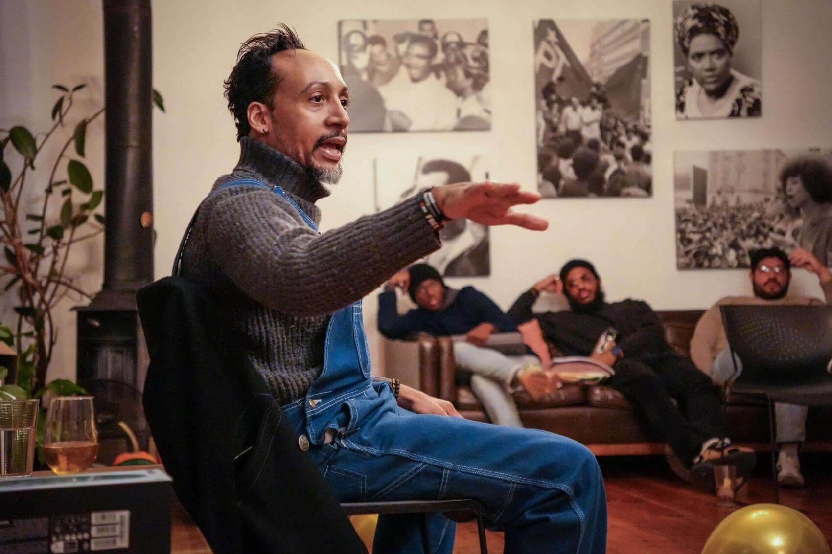 A speaker gestures with one hand while addressing a seated audience in a room with photos on the wall.