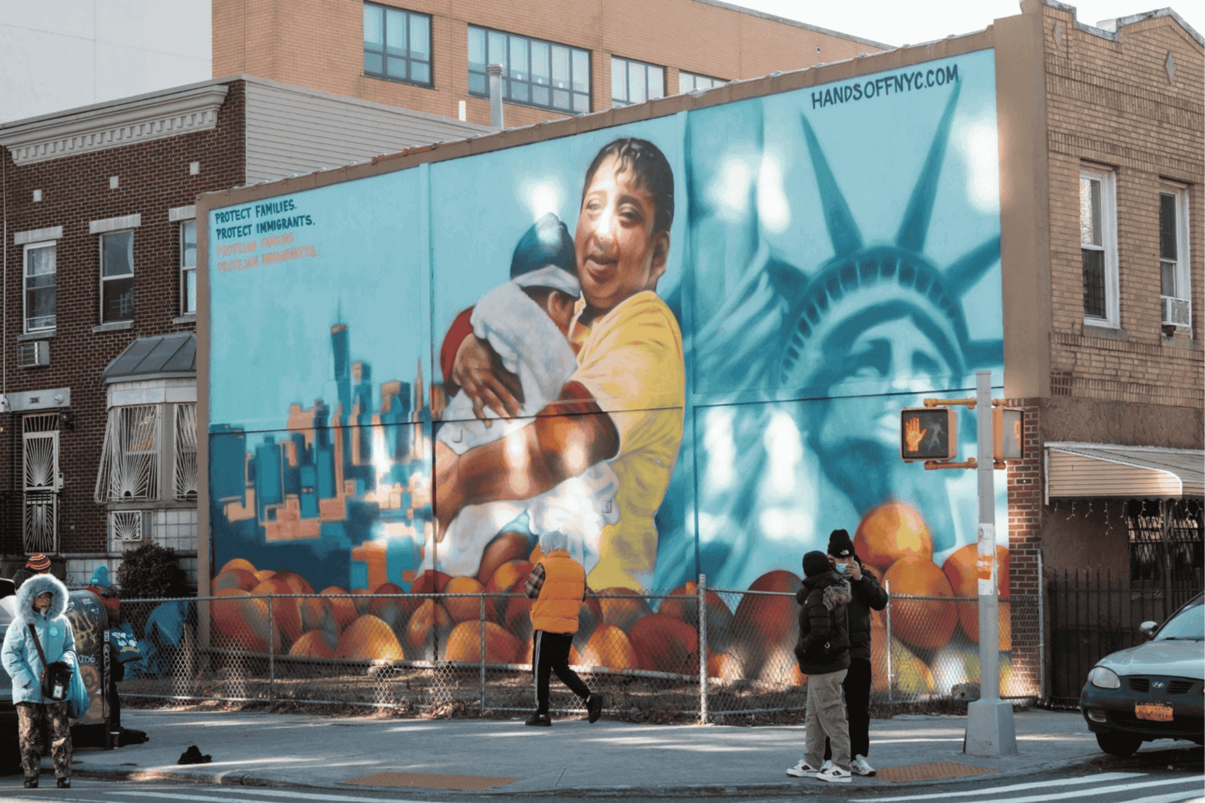 Mural of a person holding a child, with text advocating for immigrant protection, on a building in Brooklyn.