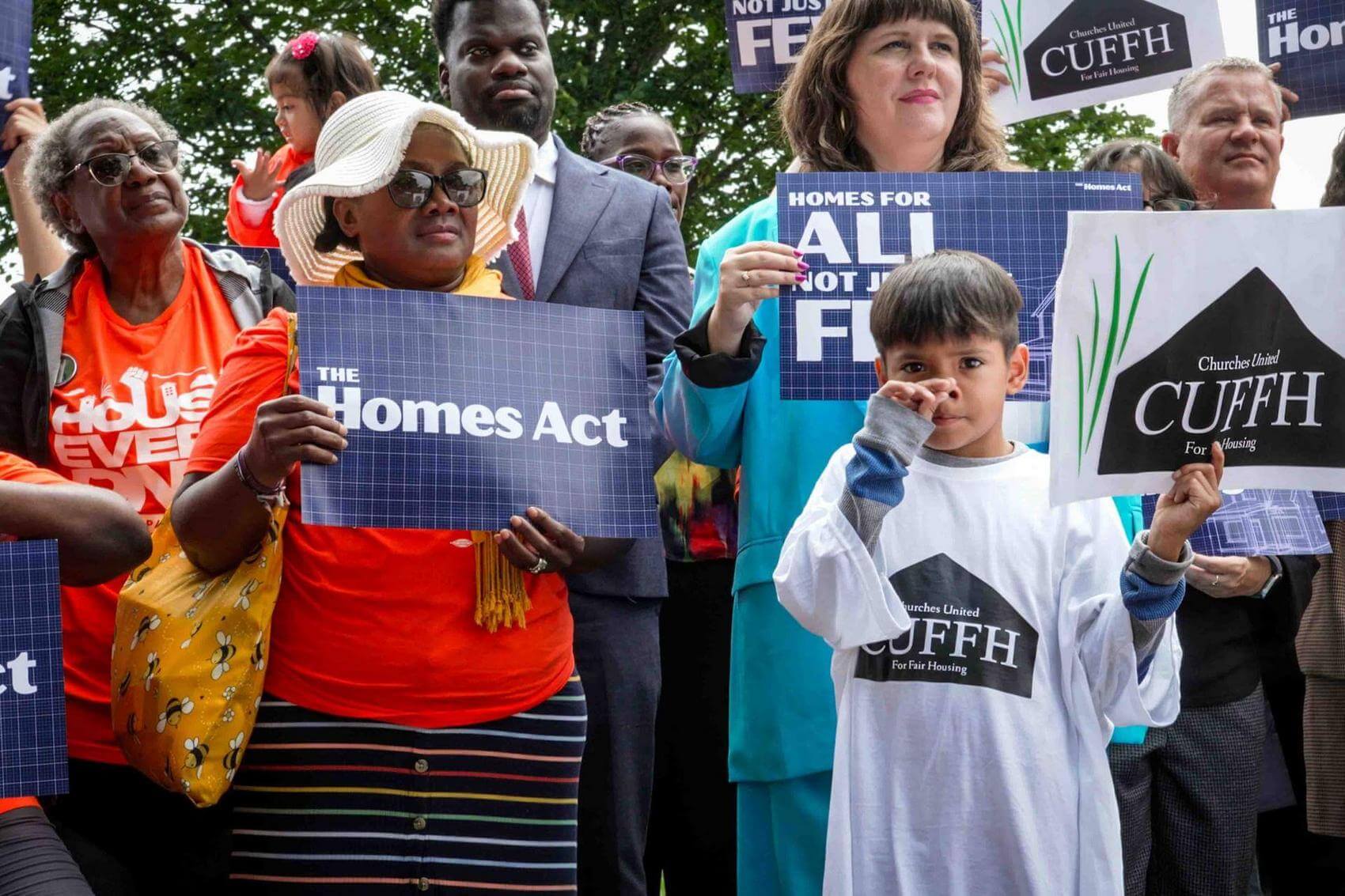 People in yellow shirts clapping and holding signs at a rally, with cameras in the foreground.