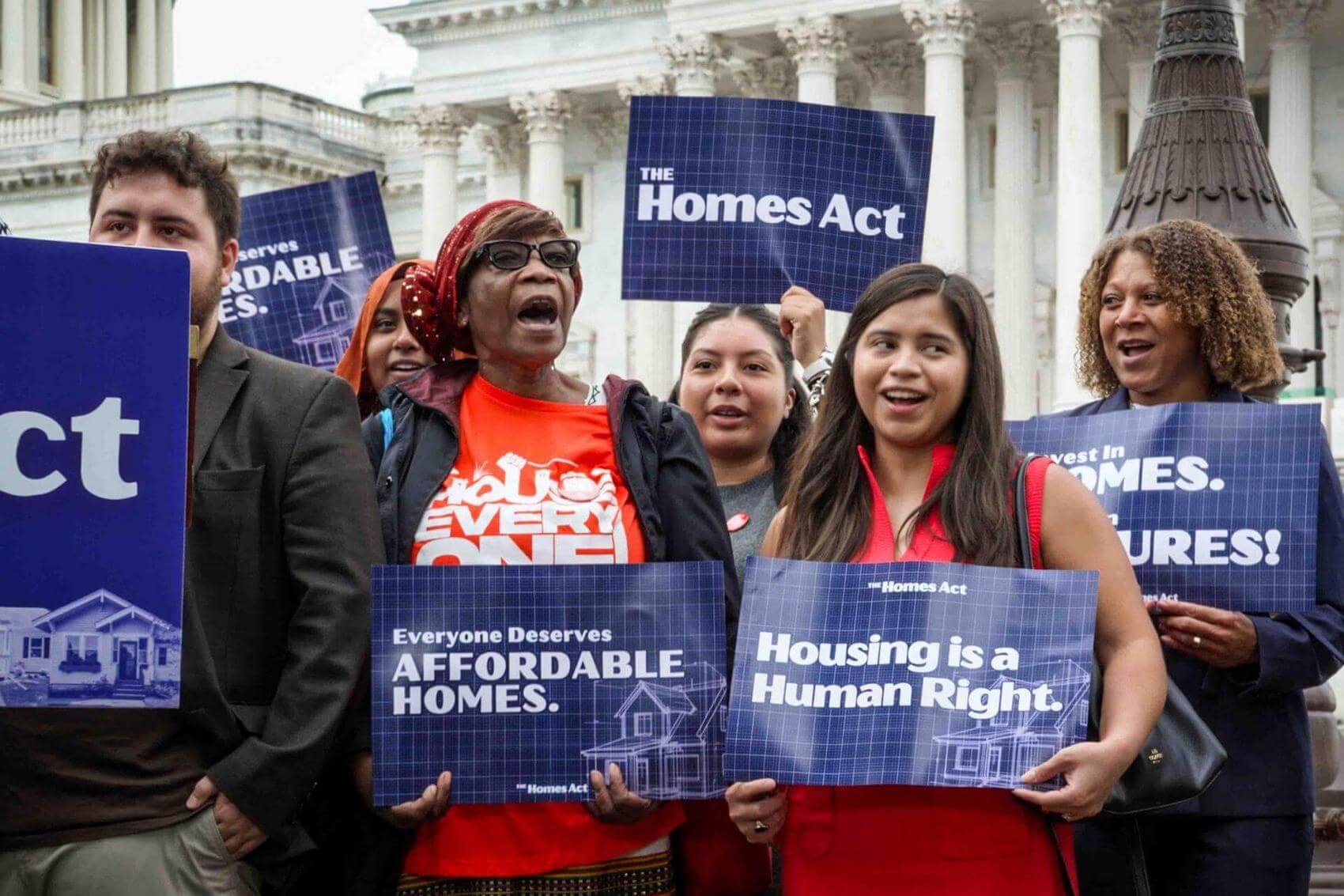 Politician speaking at podium with sign "The Homes Act" in front of Capitol building, crowd holding signs supporting housing rights.