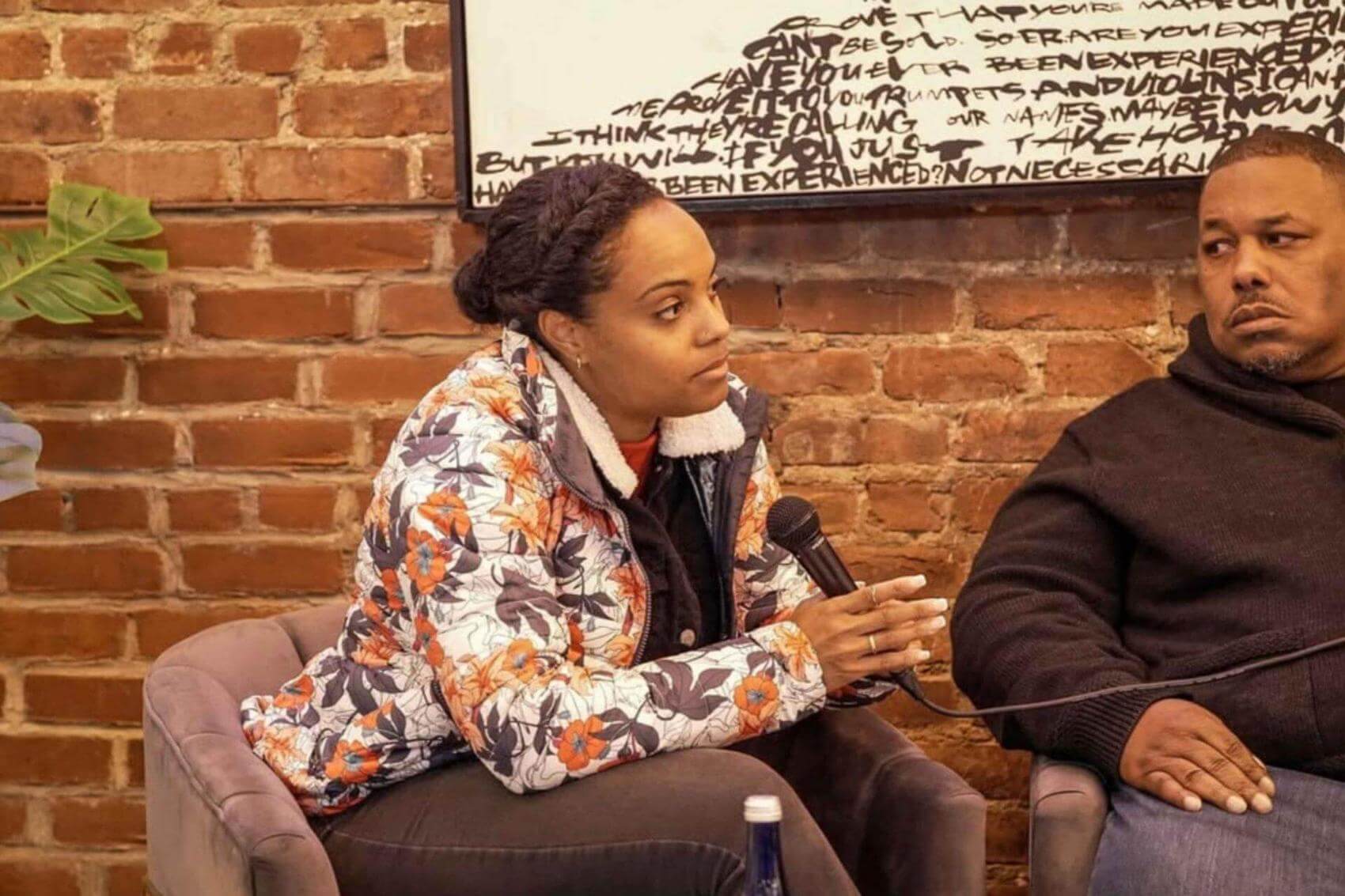 Three men sit in chairs facing forward in a room with brick walls, while a woman stands nearby.