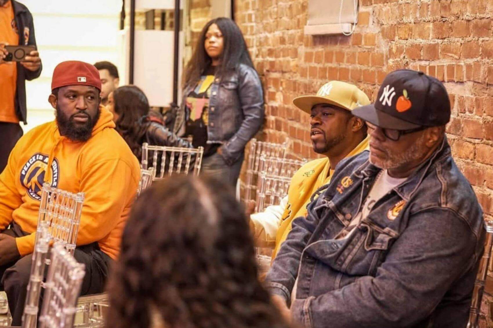 Three people sitting in transparent chairs in a brick-walled room.