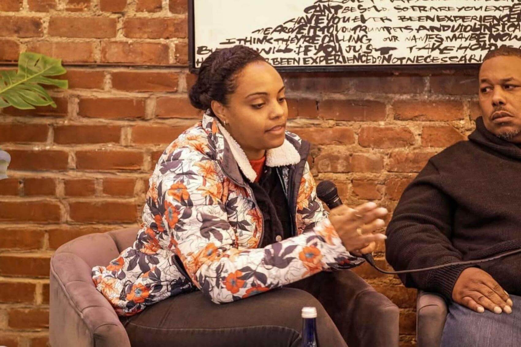 Two people seated in armchairs against a brick wall, one speaking into a microphone.