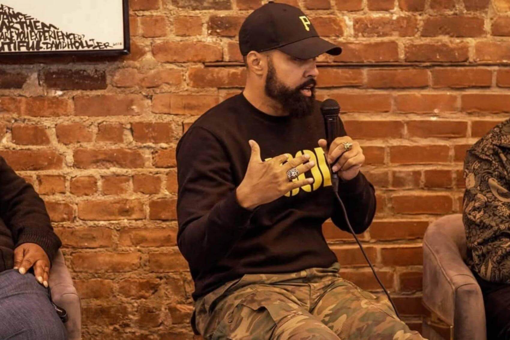 A person speaks into a microphone while sitting on a chair, with another person beside them.