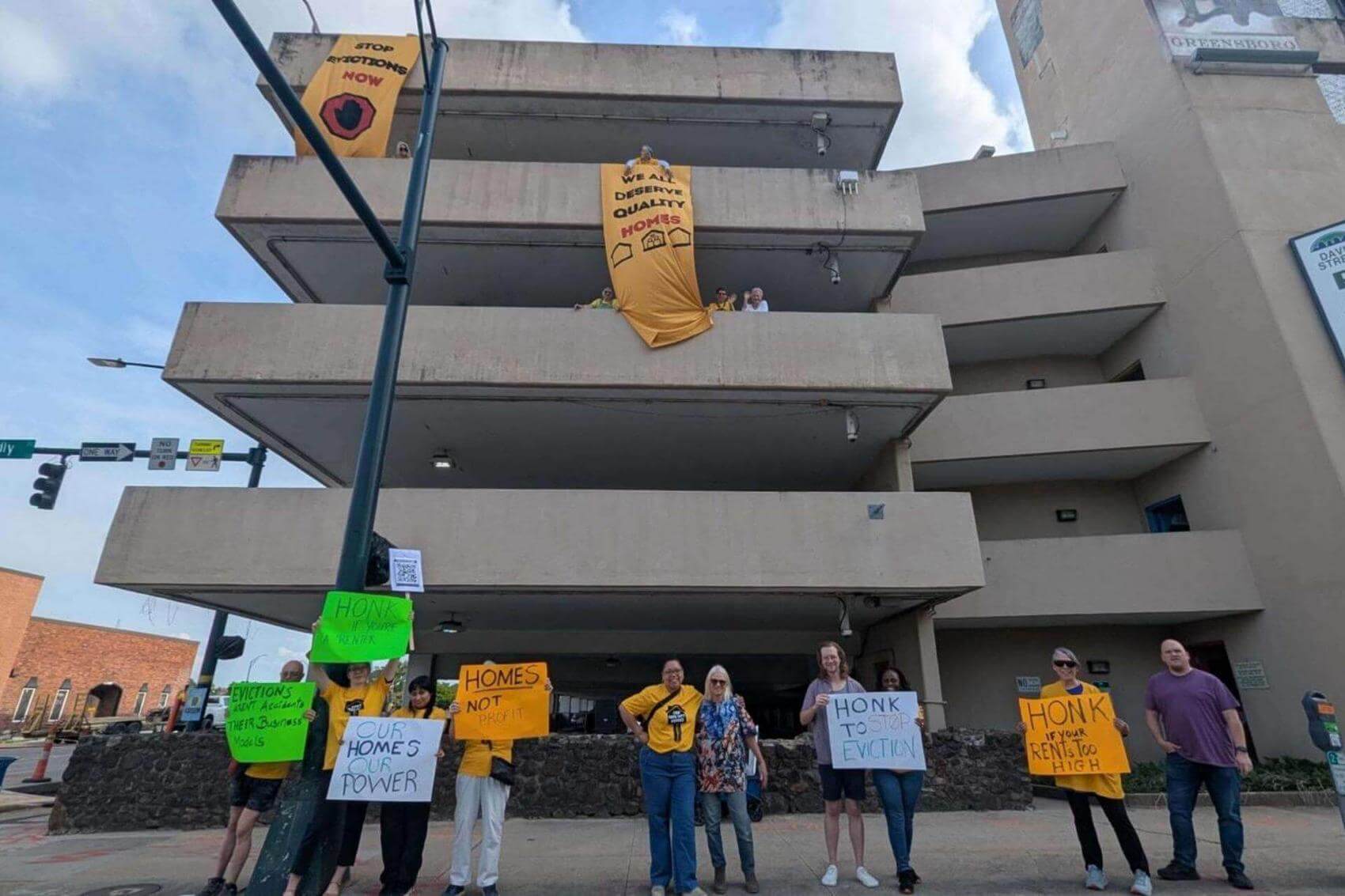 Protesters holding signs against evictions in front of a concrete parking structure.
