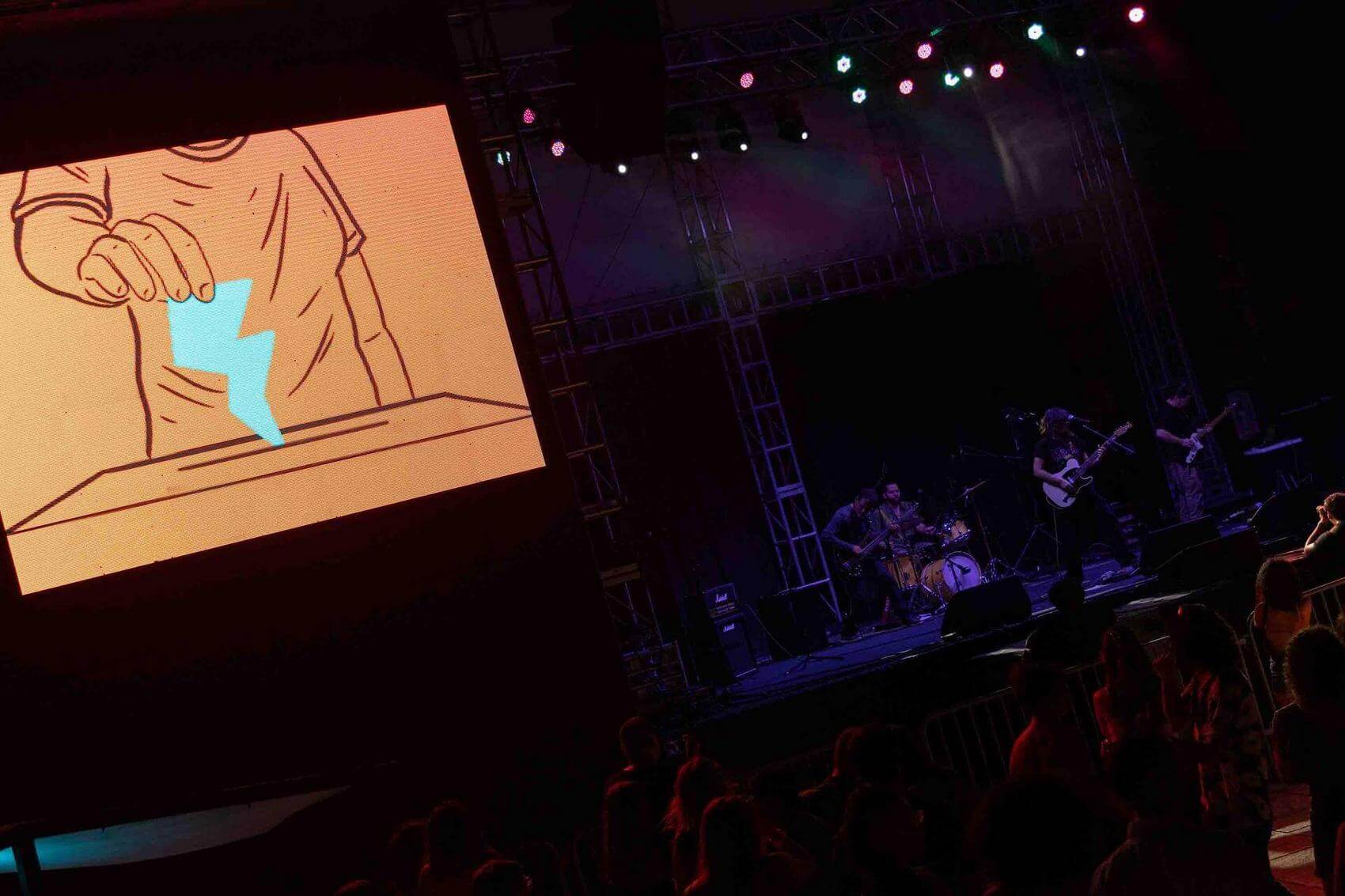 Person in colorful hat and pants speaking into microphone on stage with group holding microphones and paper.