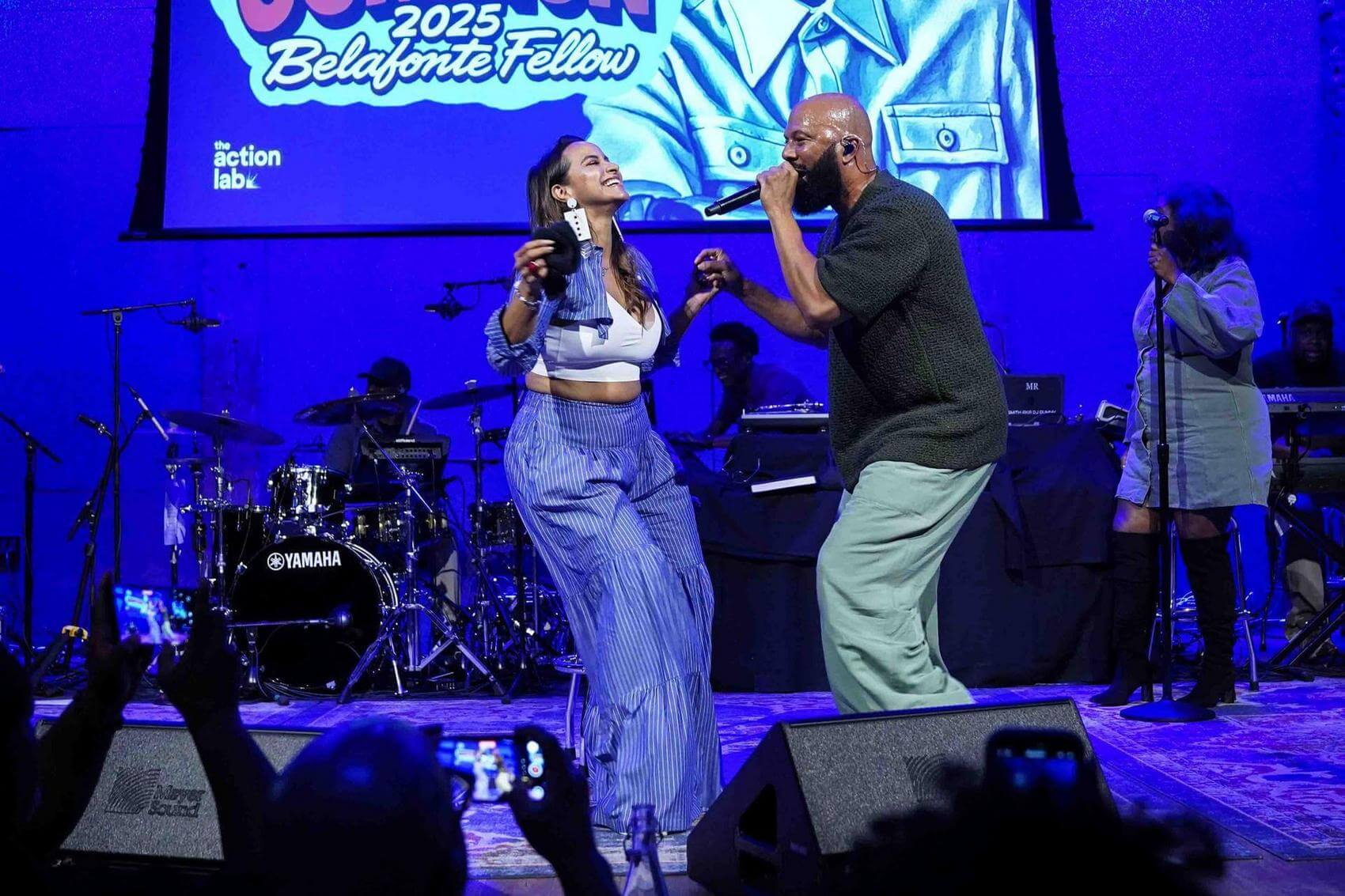 Person speaking into a microphone on stage with an audience in the background.