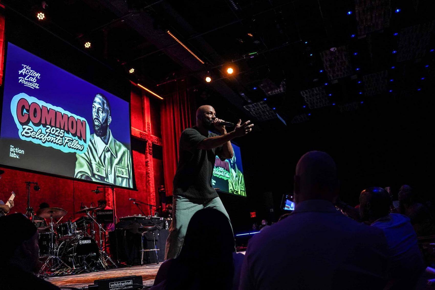 The rapper Common on stage with with arms crossed, while a woman sings into a microphone.