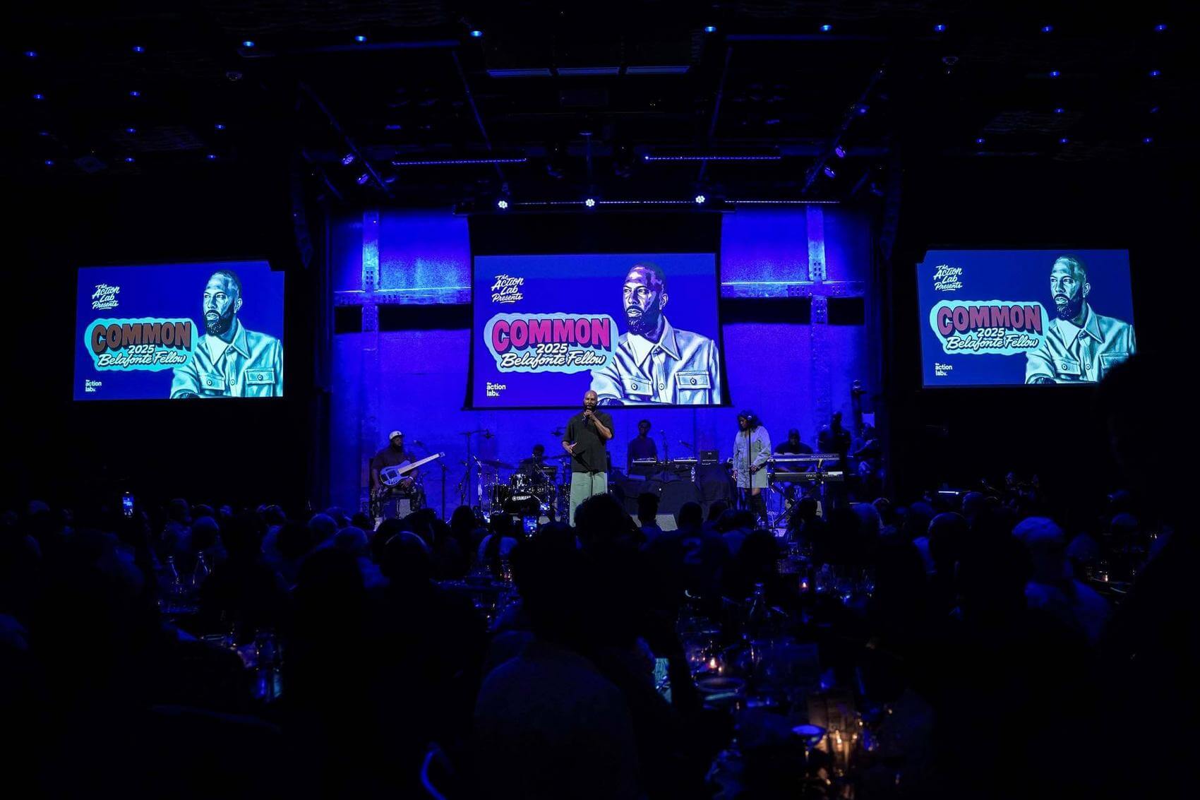 Man and woman performing on stage, with musicians and audience visible in the background.