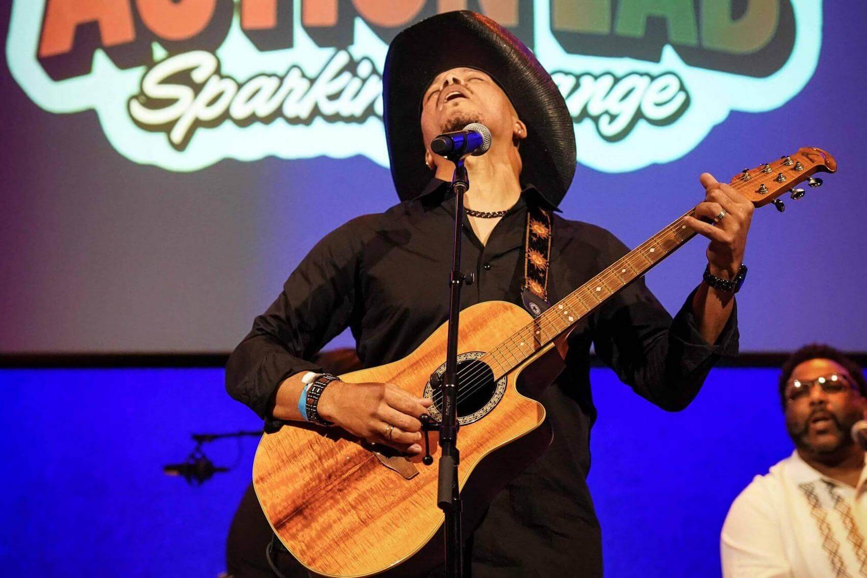 Two people pose for a photo with their arms around each other in front of a music venue poster.