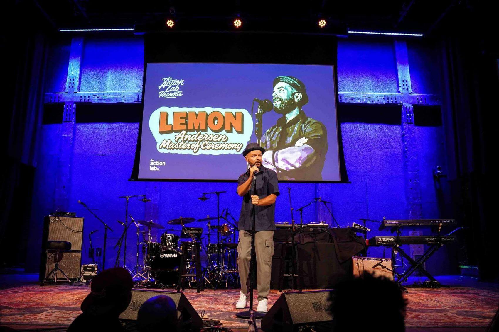 Person singing into a microphone on stage with a digital backdrop showing "Ana Tijoux 2023 Belafonte Fellow."