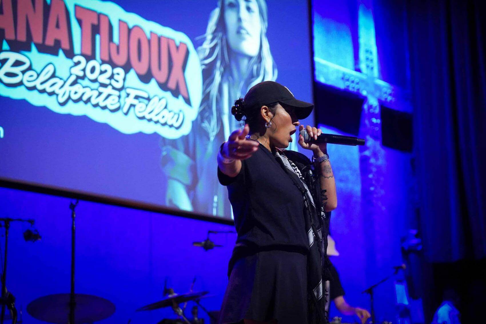 Artist performing on stage with a microphone, crouching, and engaging the audience.