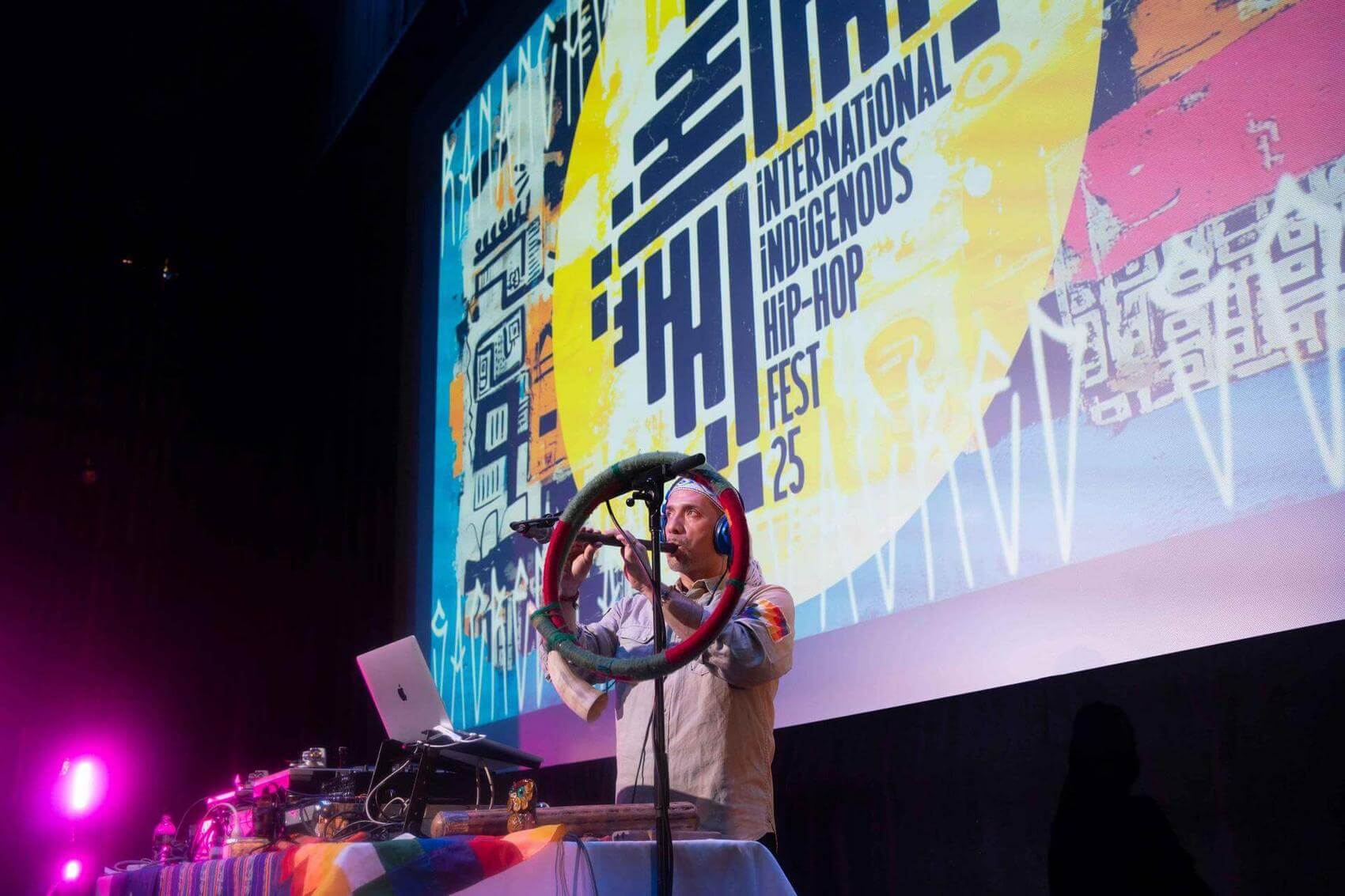 A performer plays a flute while standing in front of a colorful backdrop for a hip-hop festival.