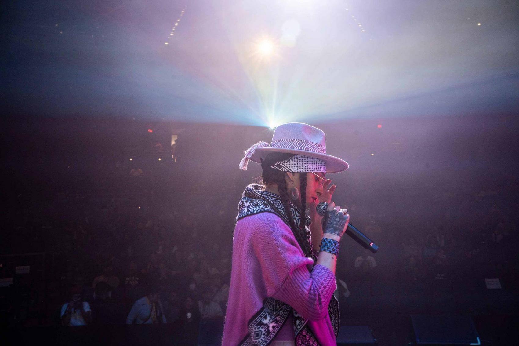 A performer raps into a microphone with one hand raised, while a DJ plays music behind.