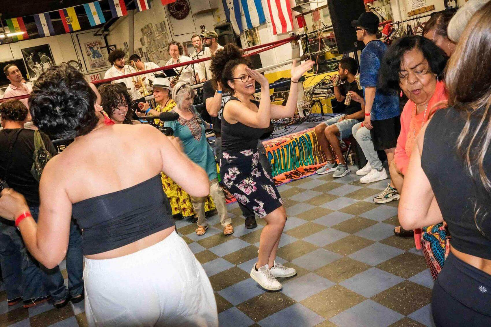 A group of eight people seated on a bench in front of a colorful mural and flags.