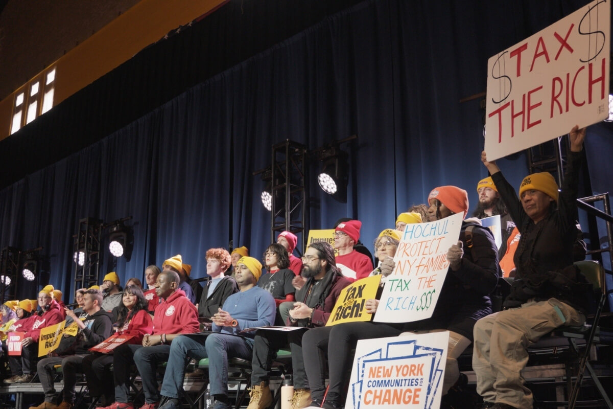 People seated on stage holding signs with messages about taxing the rich.