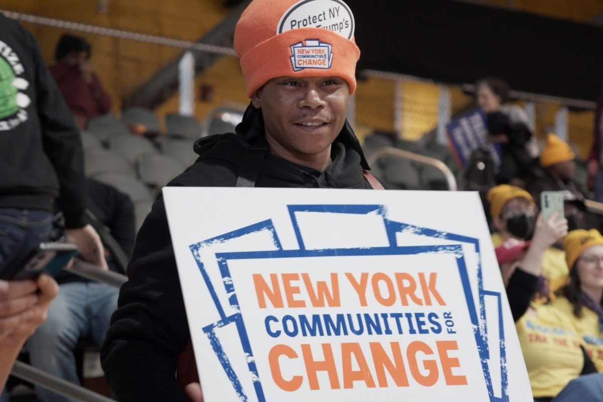 Person wearing orange hat holding a sign reading "New York Communities for Change"