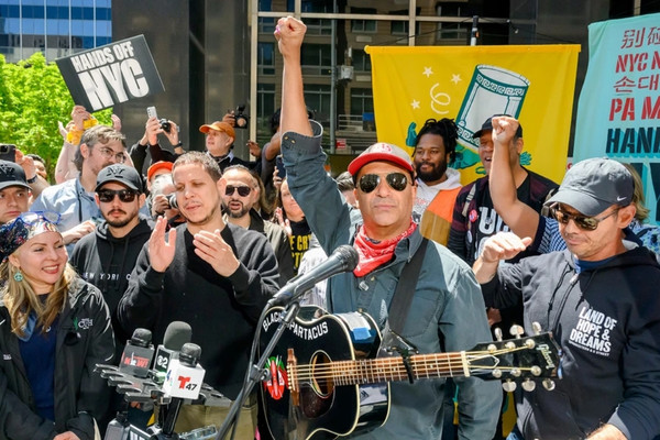 Tom Morello performs at an anti-ICE rally in New York City surrounded by protesters, organizers, and signs demanding protections for immigrant communities.