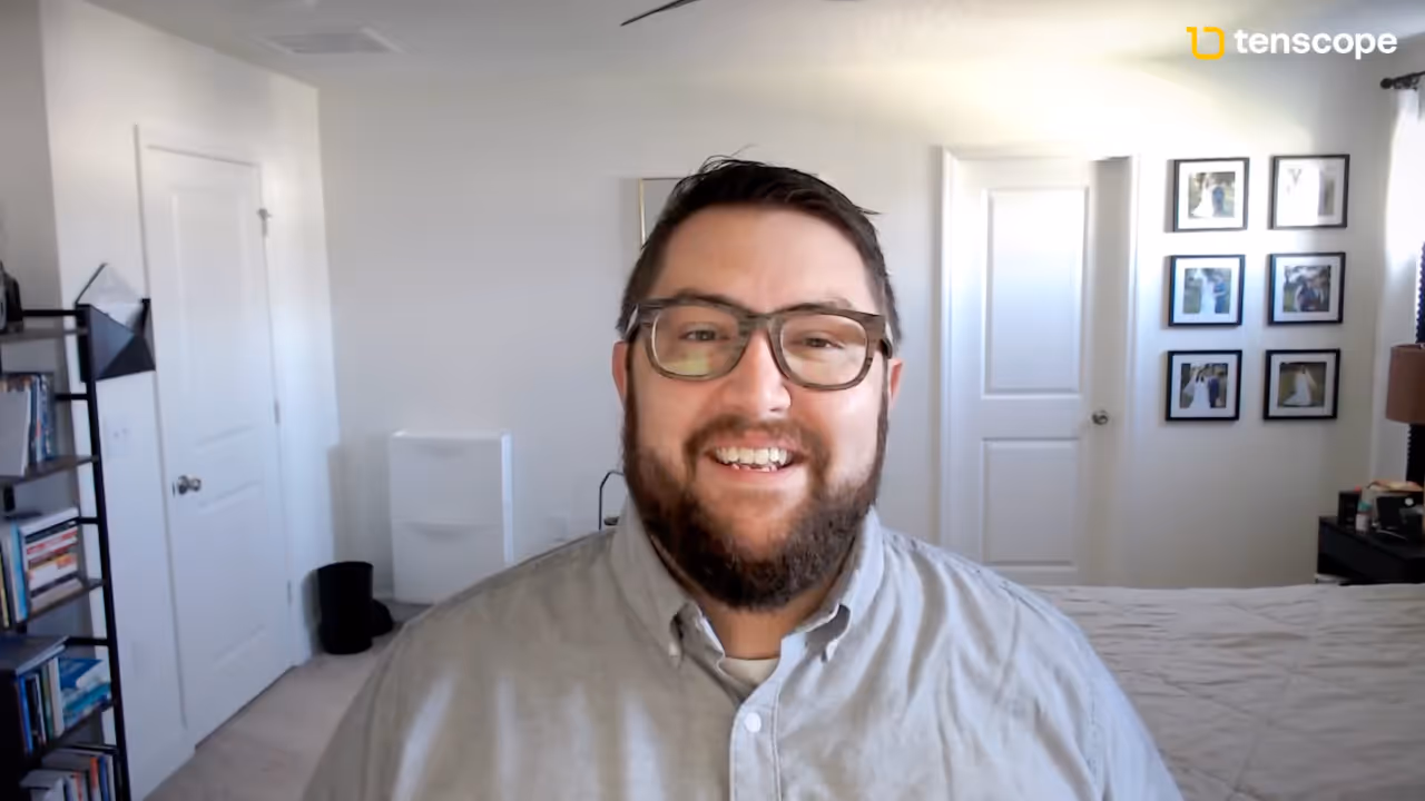 Bearded man wearing glasses and a light grey button-up shirt smiling in a well-lit bedroom with white walls and framed photos.
