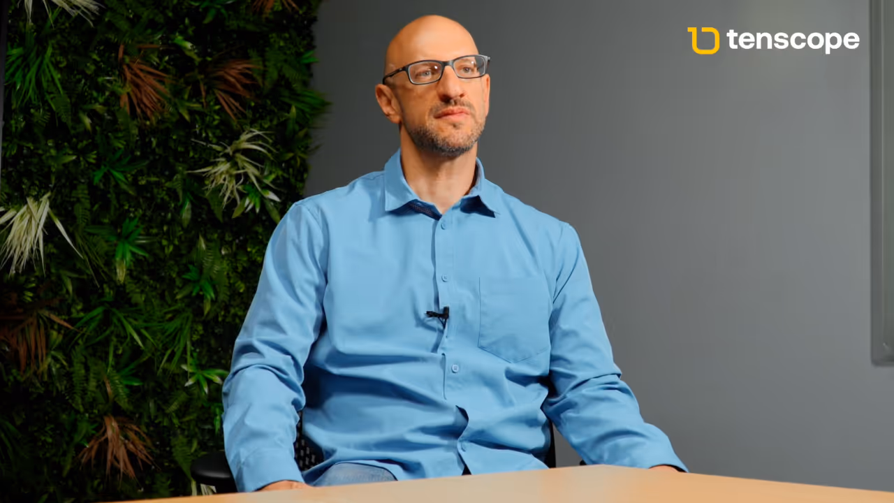 Bald man with glasses wearing a light blue shirt sitting at a table with a green leafy background and gray wall with Tenscope logo.