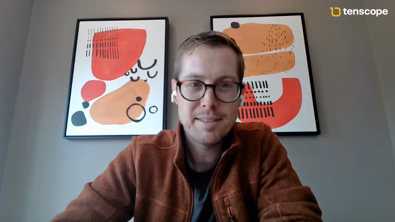 Man wearing glasses and a brown jacket sitting in front of two abstract art paintings on a gray wall with a Tenscope logo in the top right corner.