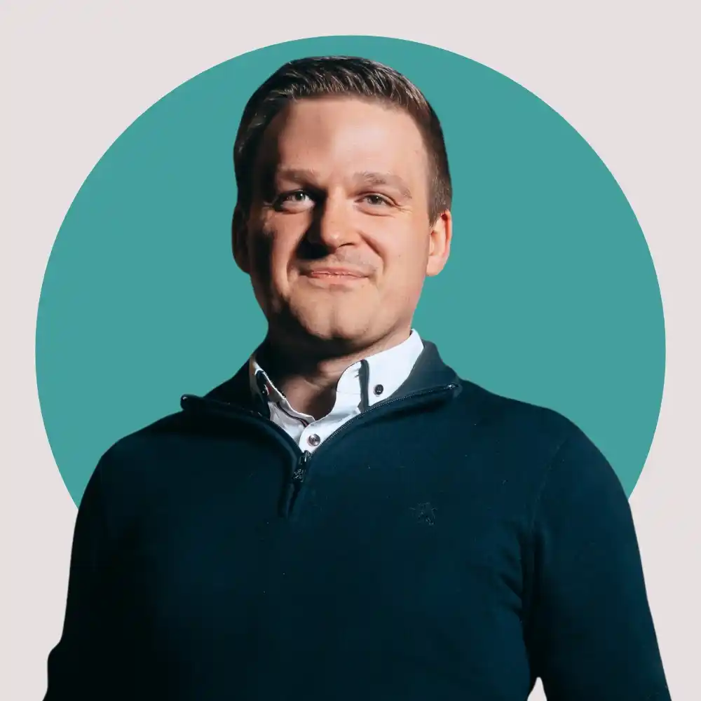 Mann mit kurzen braunen Haaren, weißem Hemd und dunklem Pullover vor türkisem Kreis auf hellem Hintergrund.