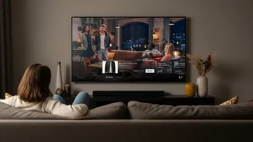 Woman in cream sweater sitting on a couch watching a smart TV with an interactive shopping interface displaying a leather jacket and other apparel in a modern living room.