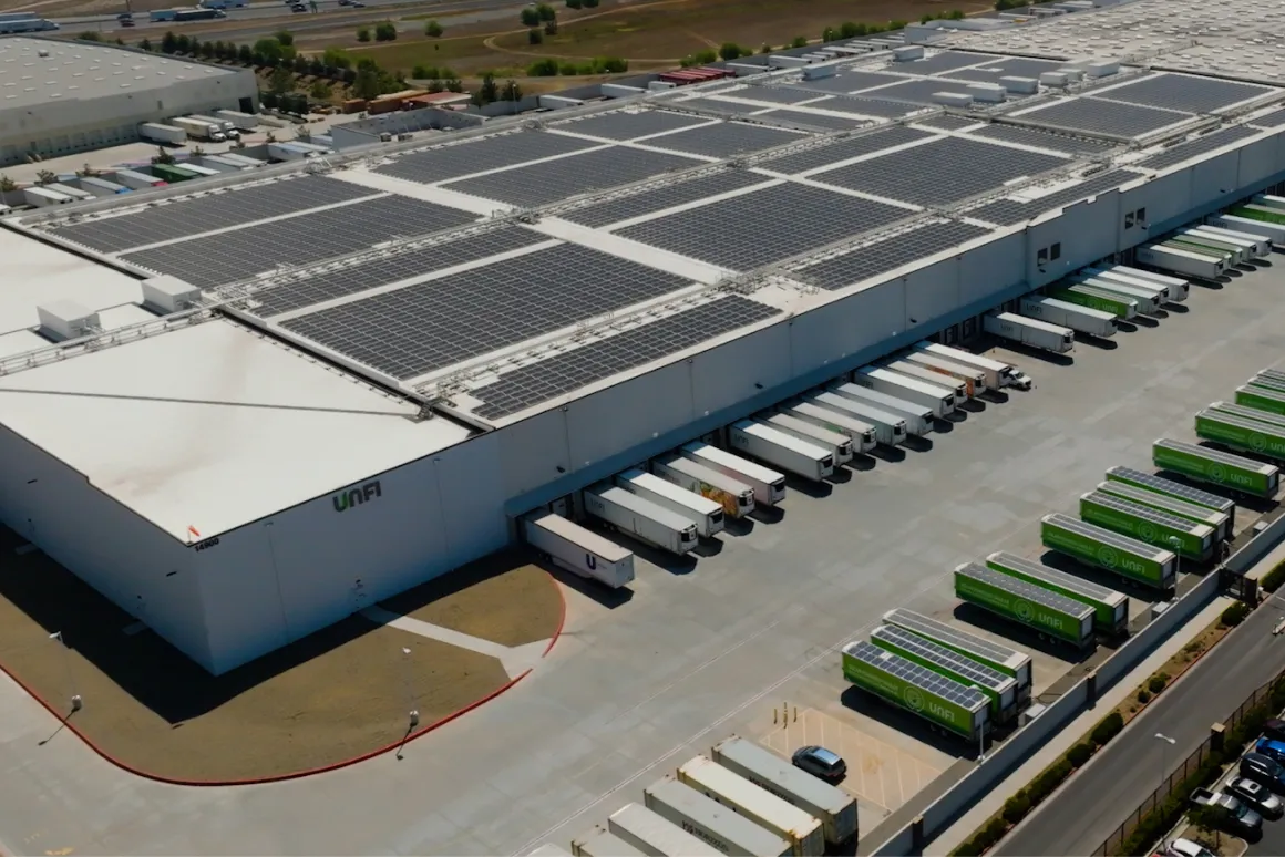 Aerial view of a large UNFI warehouse with solar panels on the roof and delivery trucks parked at loading docks.