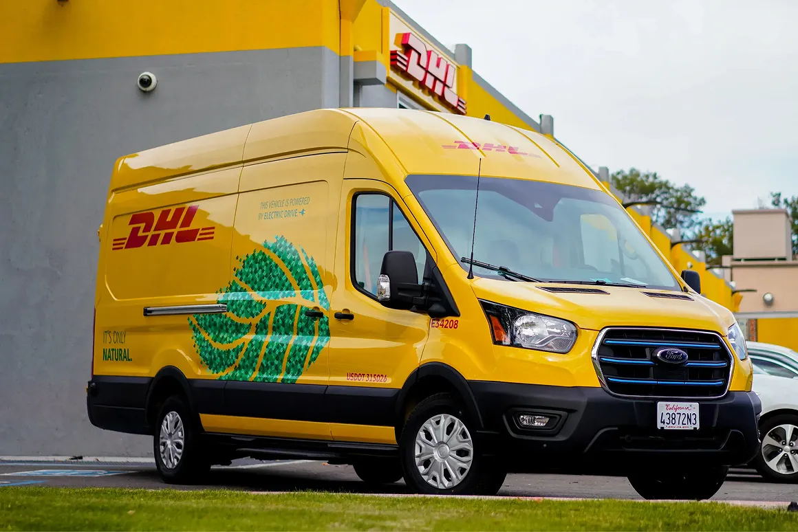 Yellow DHL delivery van with green leaf design and text indicating it is powered by electric drive, parked in front of a DHL building.
