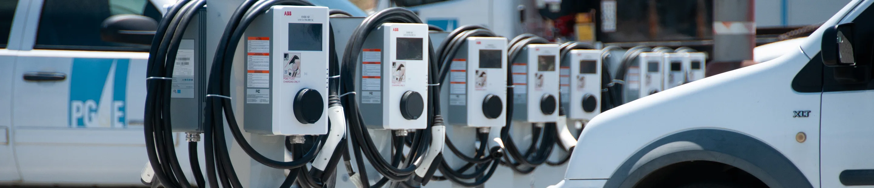 Row of electric vehicle charging stations with multiple charging cables and two white fleet trucks parked nearby.